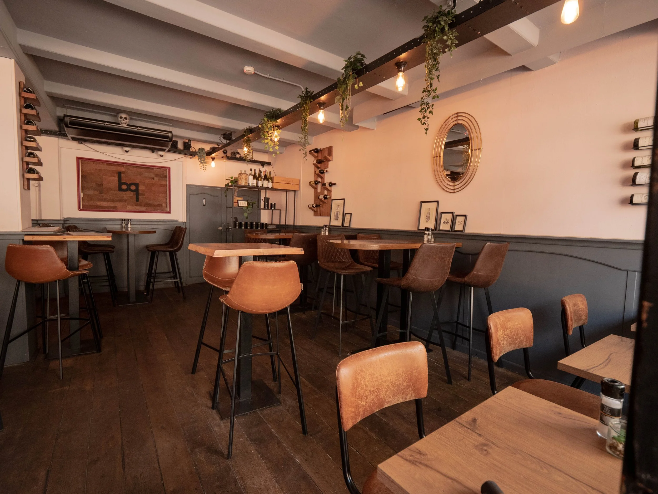 Interior de un restaurante con mesas y sillas de madera de estilo moderno, paredes decoradas con cuadros y estantes con botellas, iluminación cálida con focos y plantas decorativas.