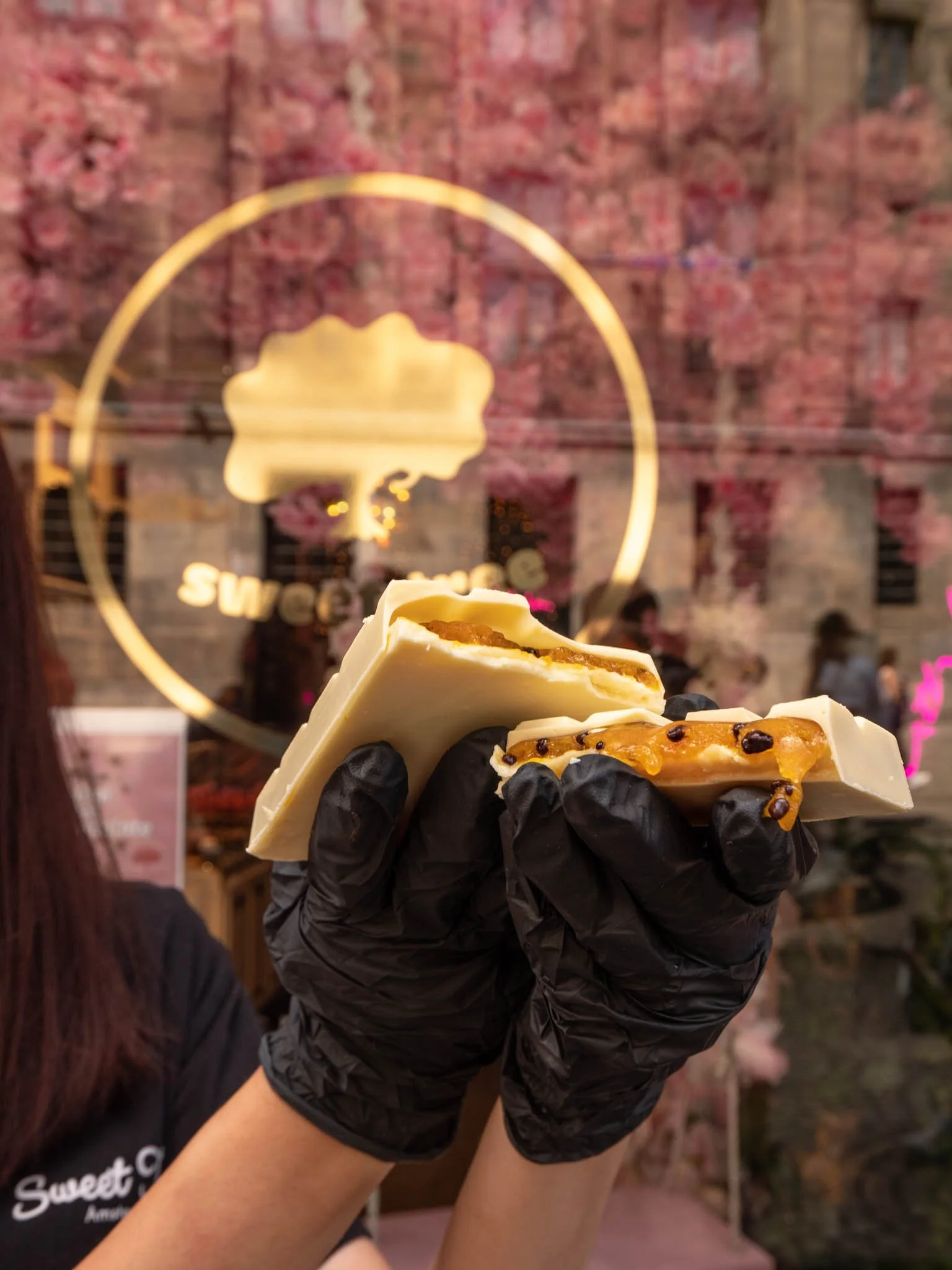 Persona sosteniendo chocolates blancos con relleno, usando guantes negros, frente a una ventana con un logo y fondo de flores rosas.