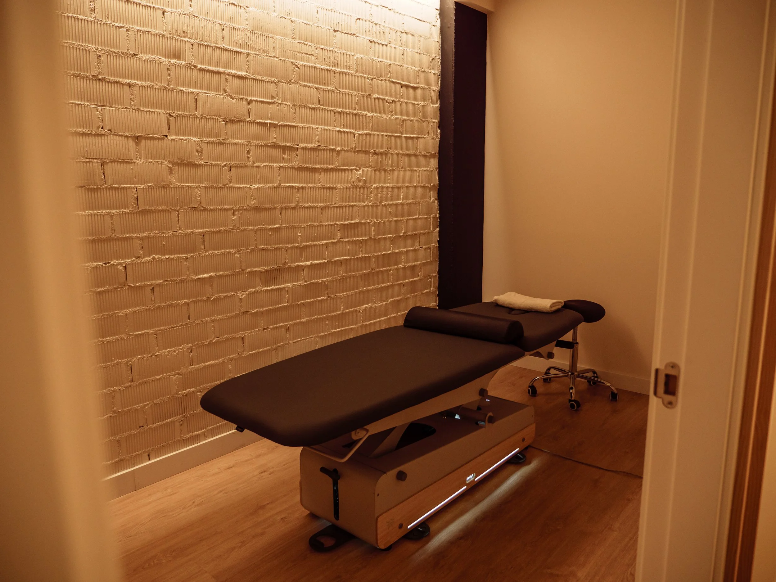 Una camilla de masaje en una sala con paredes de ladrillos pintados de blanco y una pared negra, con una toalla blanca doblada en la cabecera de la camilla