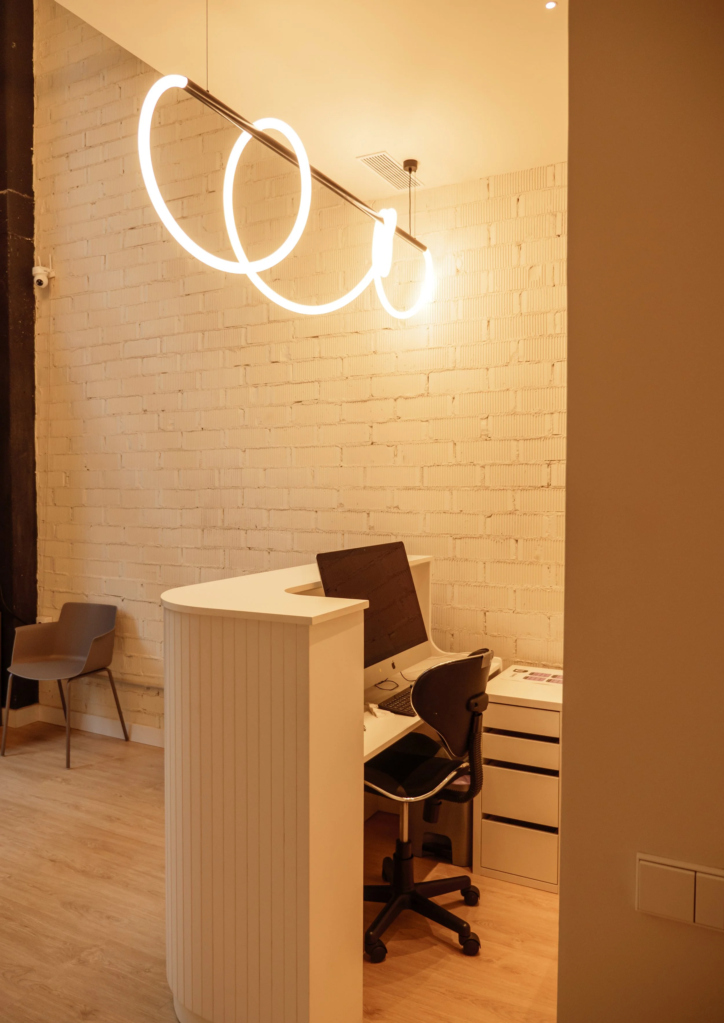 Oficina con escritorio, computadora, silla y lámpara de techo moderna en un ambiente con paredes de ladrillo blanco y piso de madera.