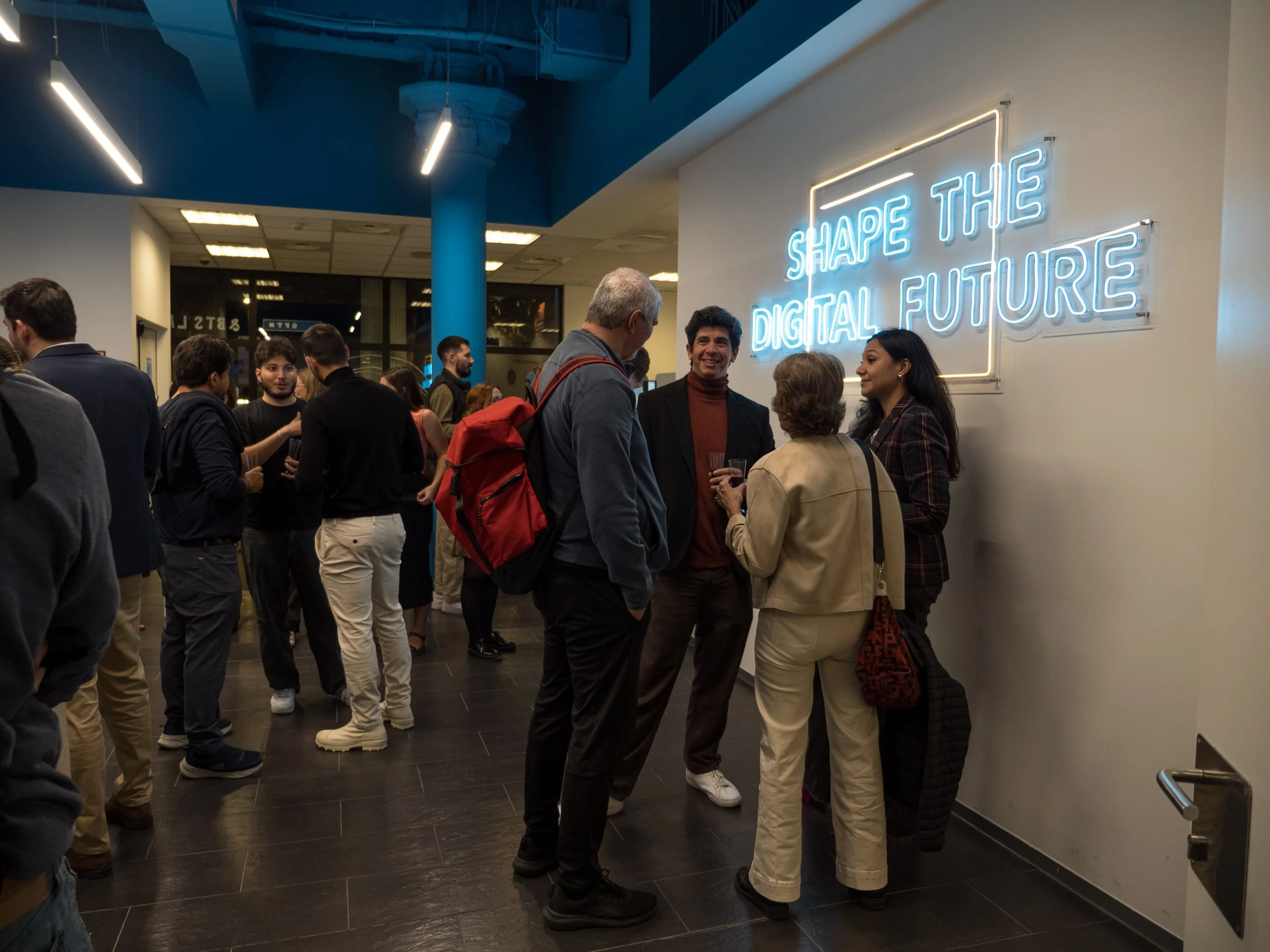 Grupo de personas conversando frente a un letrero de neón azul que dice 'Shape the Digital Future' en un evento o conferencia tecnológica.