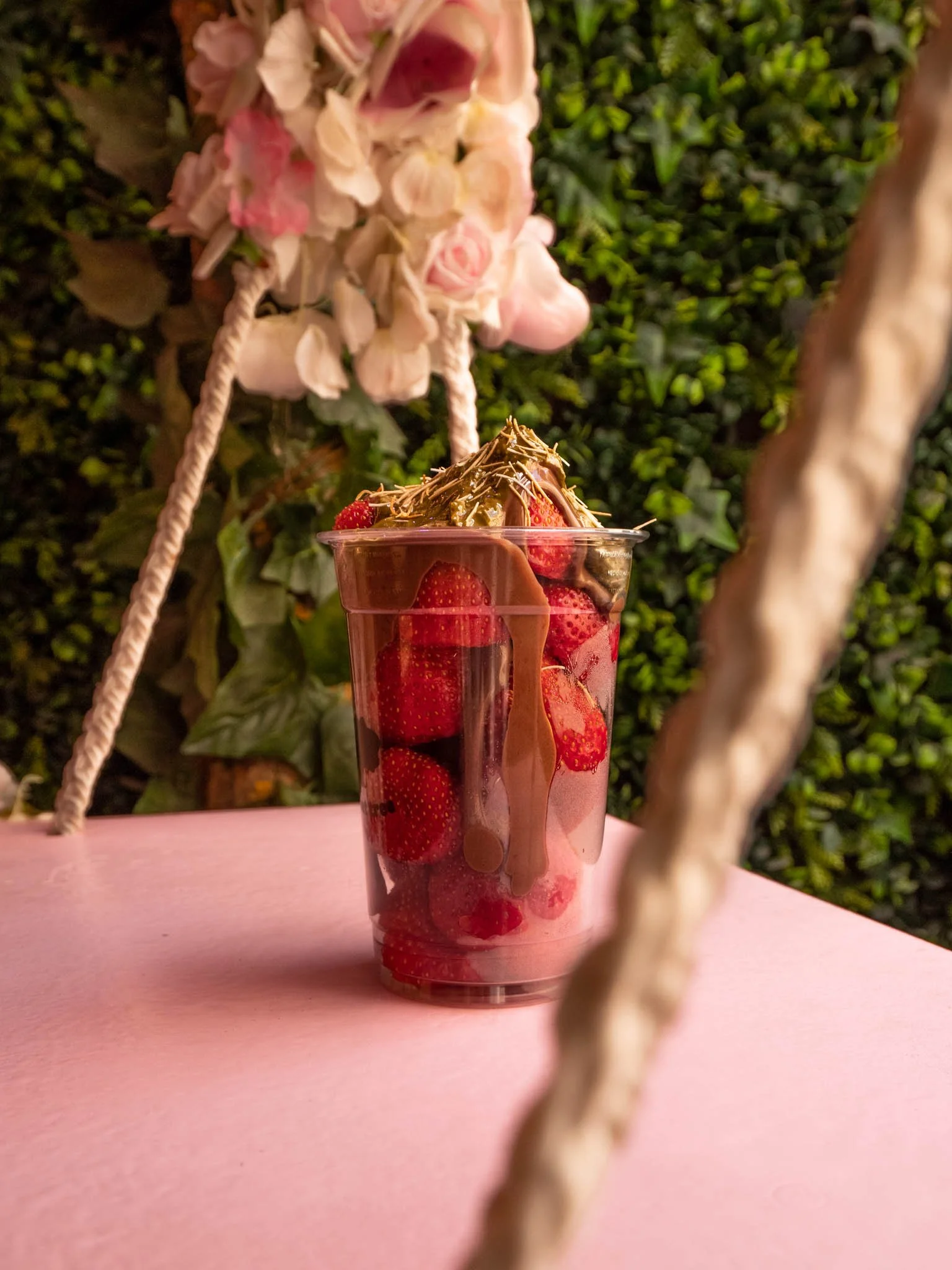 Vaso transparente con fresas y chocolate, decorado con trozos de chocolate y una ramita en la cima sobre una mesa rosa, con fondo de arbustos verdes y una estructura de cuerda con flores en la parte superior.