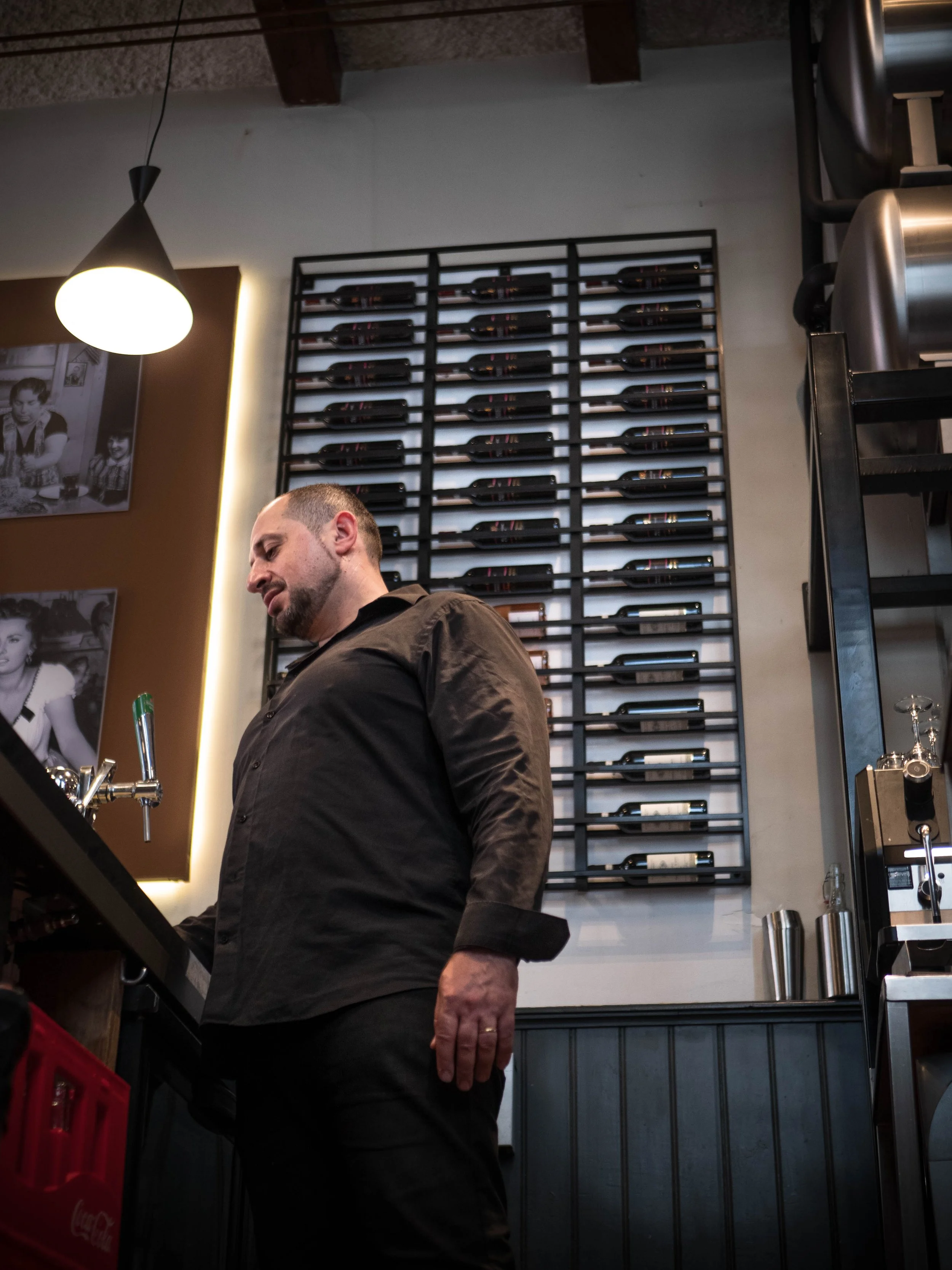 Hombre sirviendo cerveza en un bar con paredes decoradas con fotografías antiguas y estantería de botellas de vino.