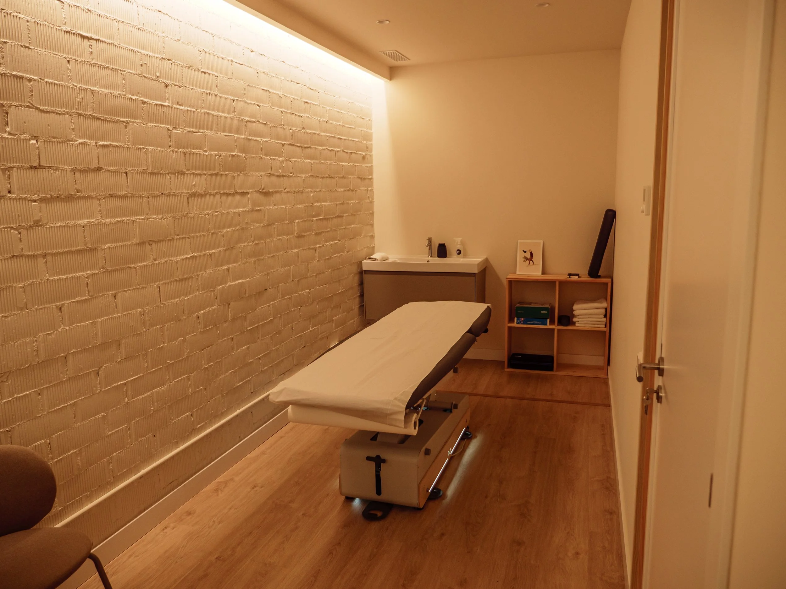Sala de masaje con camilla, pared de ladrillos blancos, estantería con toallas, y equipo de masaje en un ambiente tranquilo.