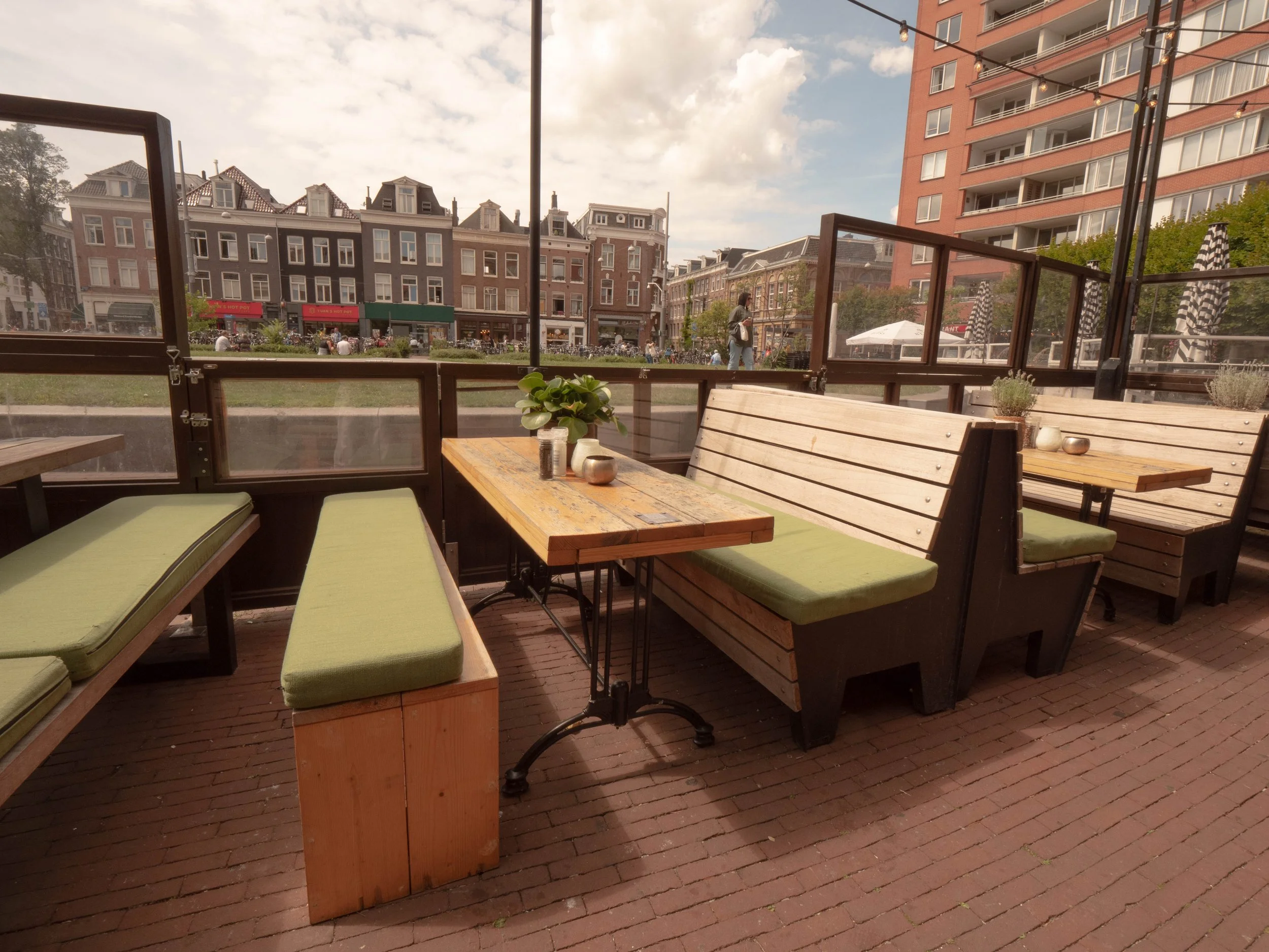 Mesa de madera con cojines verdes en una terraza al aire libre, con vista a edificios y calles con personas en un día soleado.