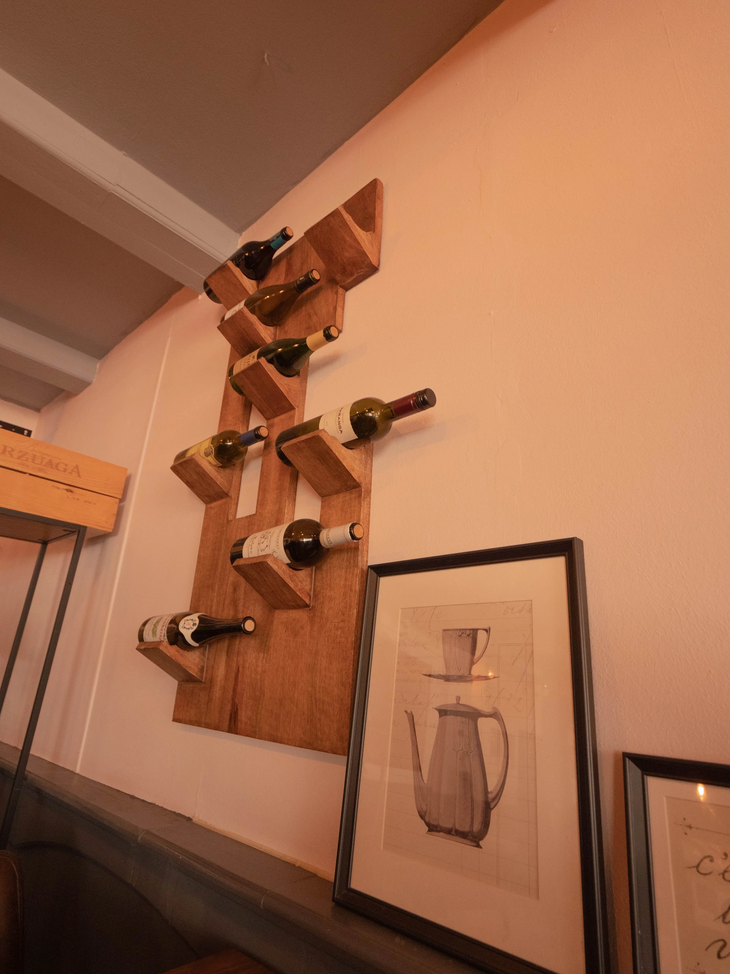 Estante de madera con botellas de vino y un marco con dibujo de una tetera y una taza.