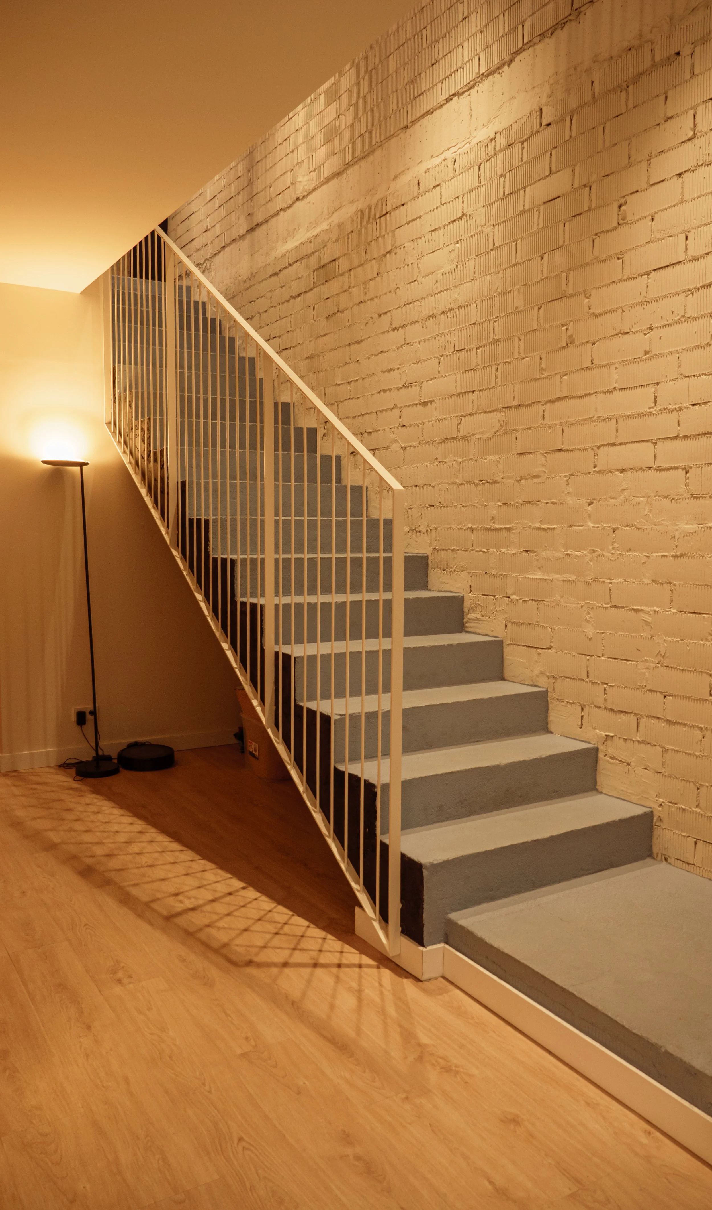 Escalera de interior con barandilla blanca, paredes de ladrillo pintadas de blanco y suelo de madera.