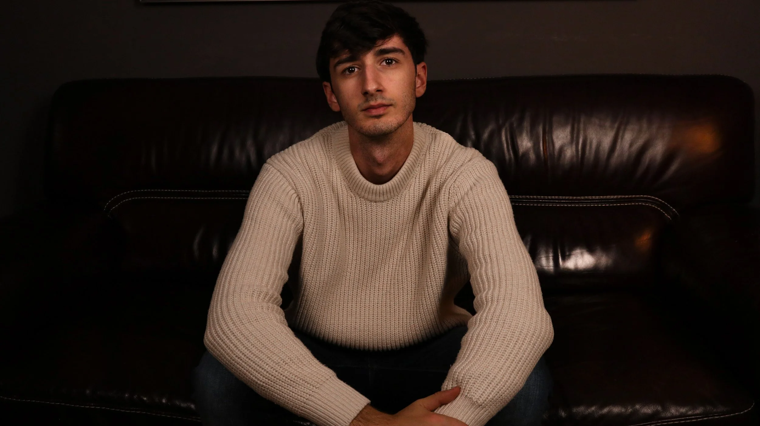 Joven sentado en un sillón de cuero oscuro, con expresión pensativa, vistiendo suéter beige de punto y barba ligera.
