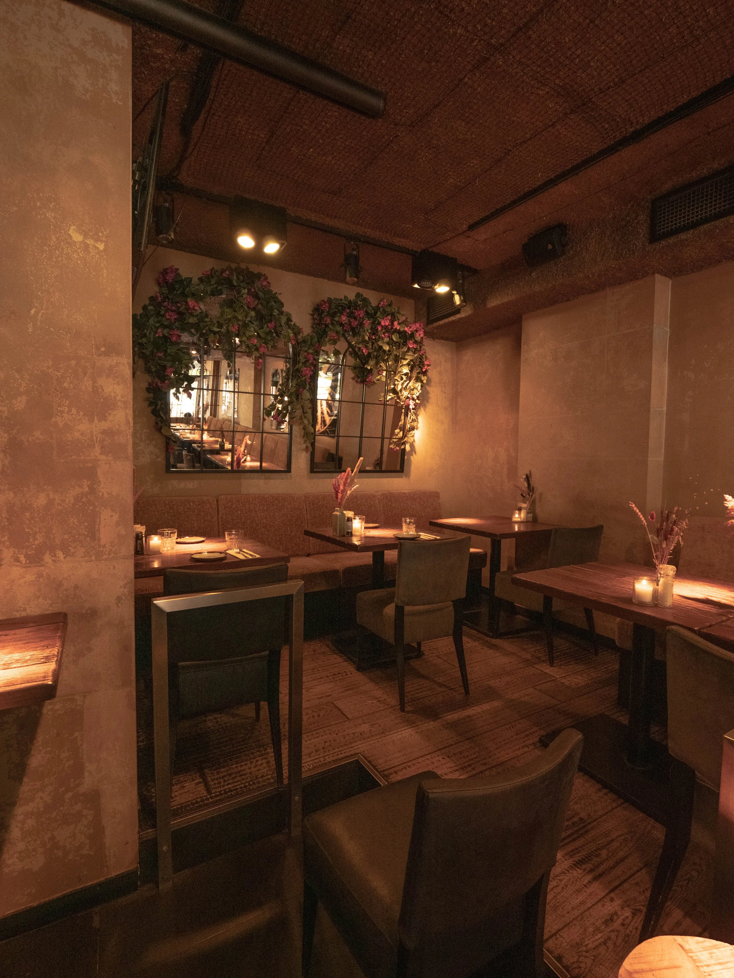 Interior de restaurante con mesas de madera, sillas tapizadas y decoraciones florales en las paredes, iluminación cálida y ambiente íntimo.