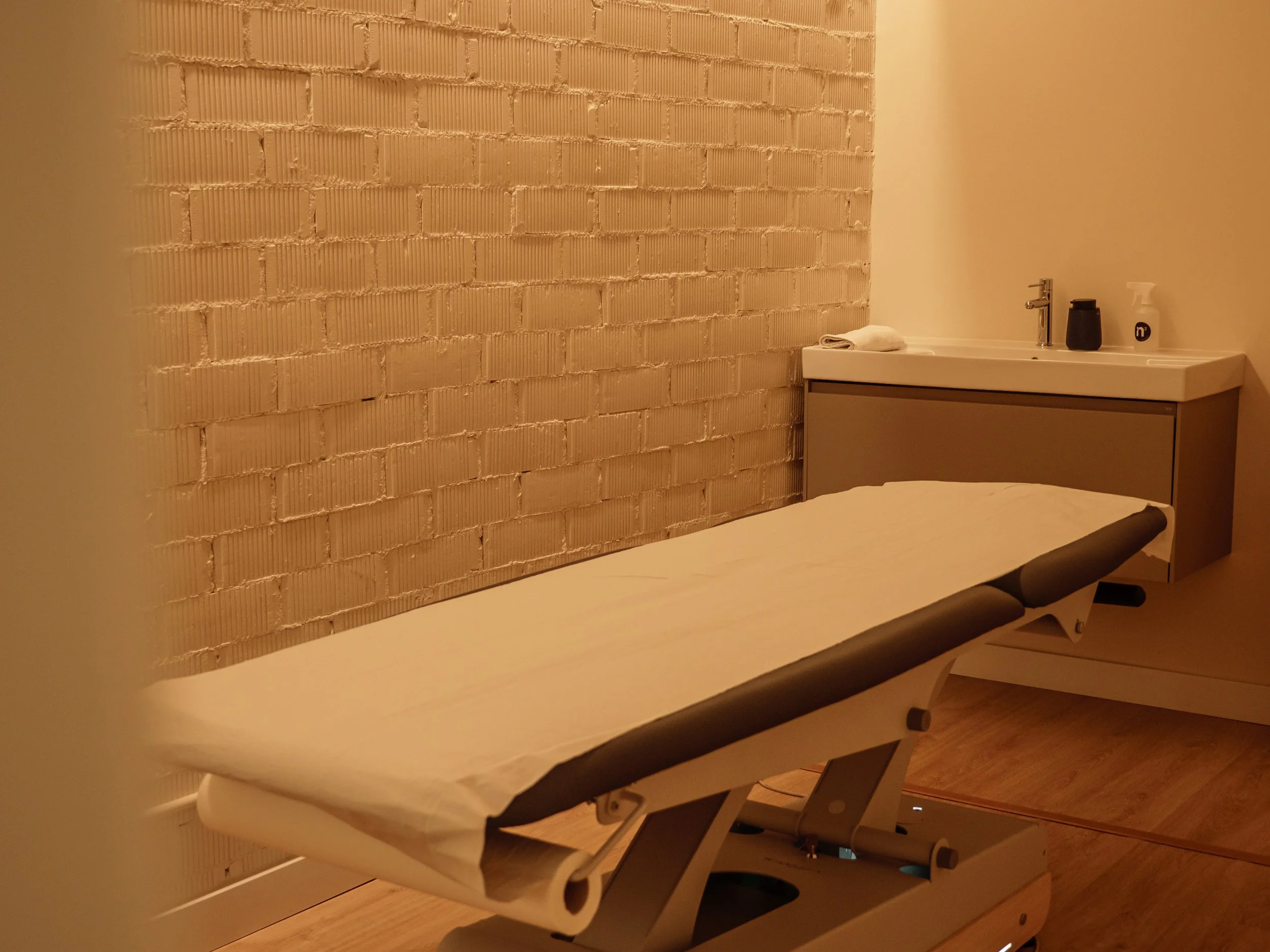 Camilla de masaje en una habitación con pared de ladrillos vista, lavabo con jabón y toalla al fondo.
