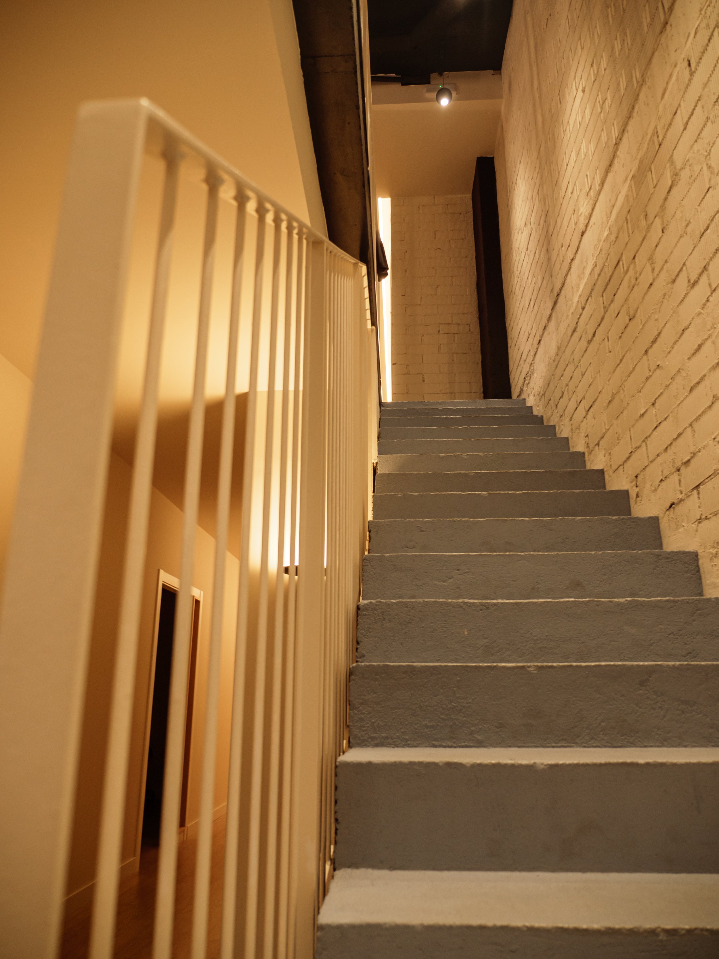 Escalera de cemento gris con barandal blanco, paredes de ladrillo pintadas de color crema, iluminación cálida y techo oscuro.