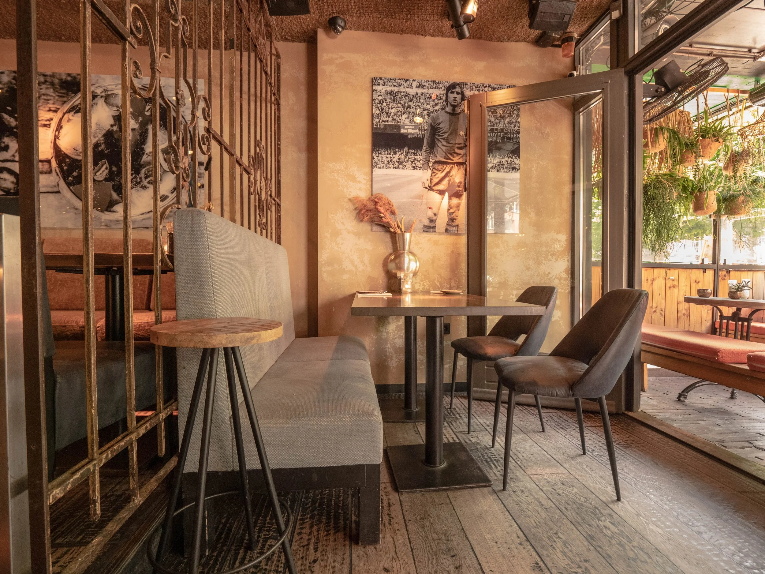 Interior de un restaurante con paredes de color beige, mesas y sillas modernas, con decoración de cuadros y plantas colgantes en la terraza exterior.