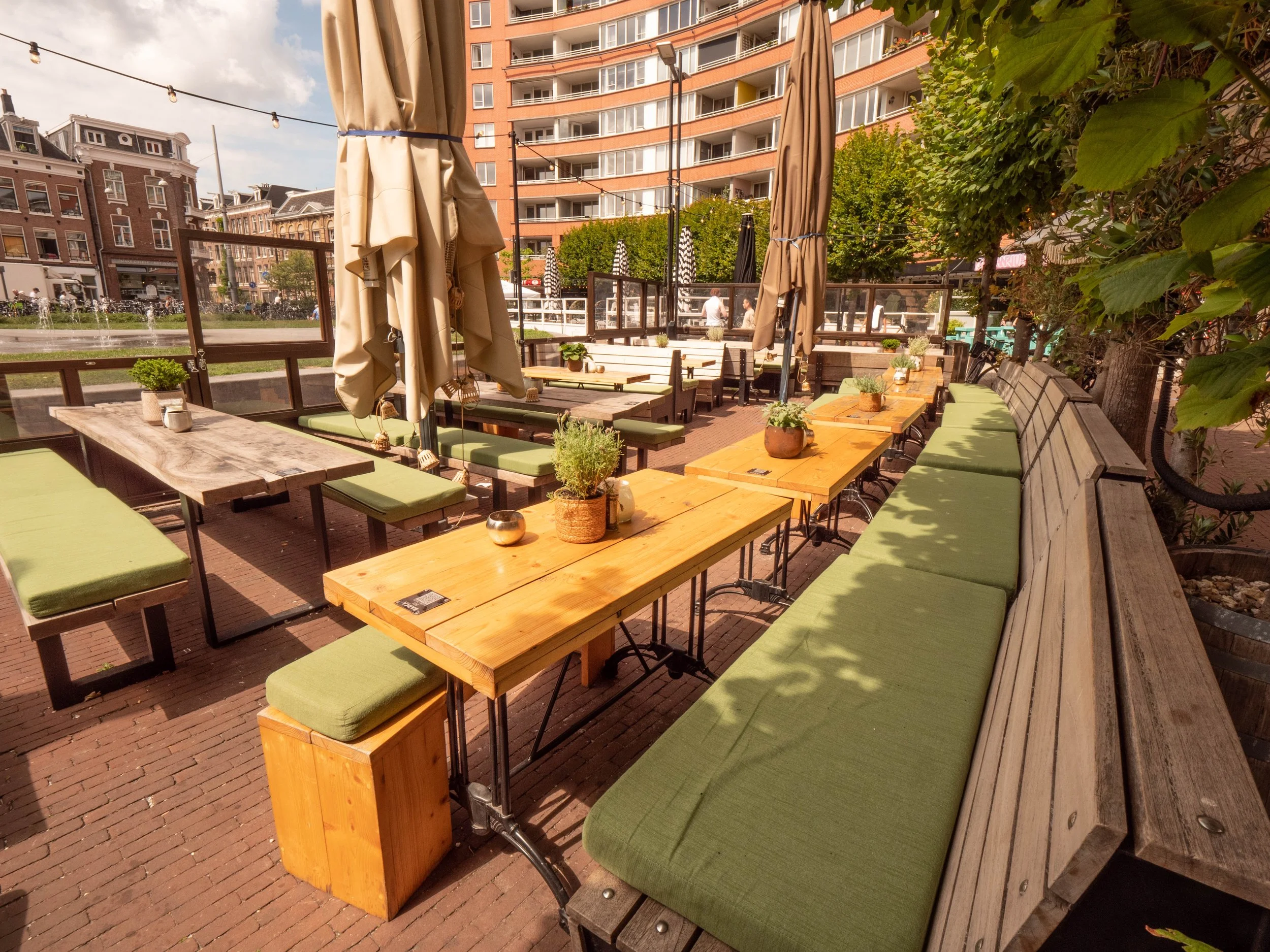 Vista de una terraza de restaurante con mesas de madera, sillas con cojines verdes, y sombrillas beige, rodeada de vegetación y edificios urbanos en el fondo.