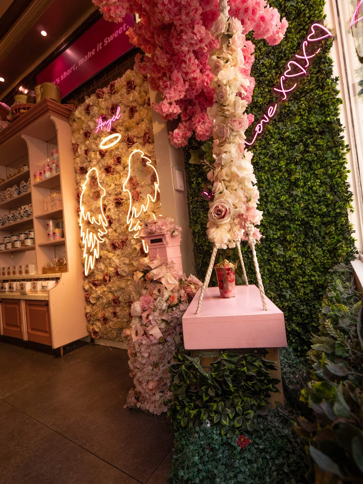 Decoración con flores en tonos rosas y blancos, con letreros de neón que dicen 'Angel' y 'Love' con corazones y alas de ángel, en un lugar acogedor con estanterías de productos y paredes cubiertas de plantas verdes.