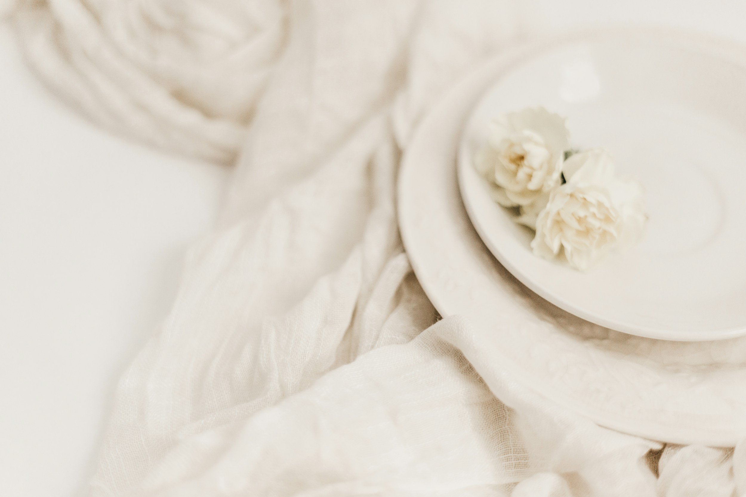 Close-up of white carnations on a white plate, surrounded by white fabric and a glass of water.