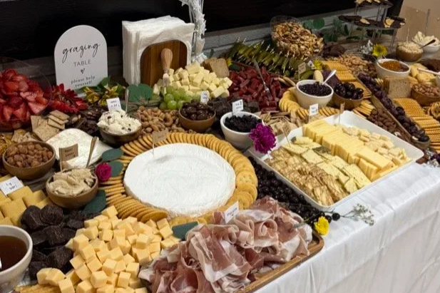 A spread of various foods on a table outdoors, including cheeses, fruits, meats, and condiments.