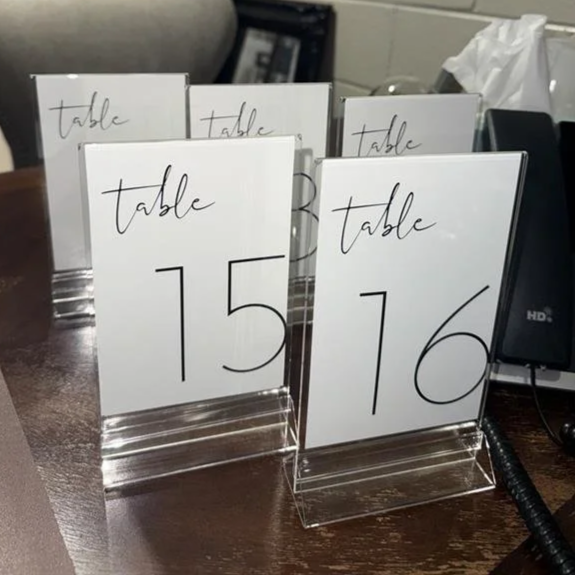 A round banquet table decorated with a floral centerpiece in a brown bottle, surrounded by multiple empty champagne glasses, small candles, and a table sign indicating it is for table 5 in a formal setting.