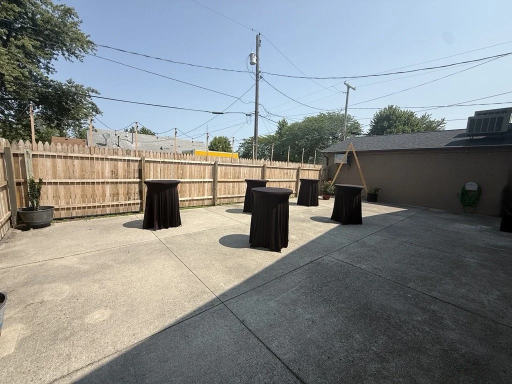 Outdoor patio with high-top tables draped in white cloths, string lights overhead, wooden fence on the left, building on the right, and a large tree in the background under a clear blue sky.