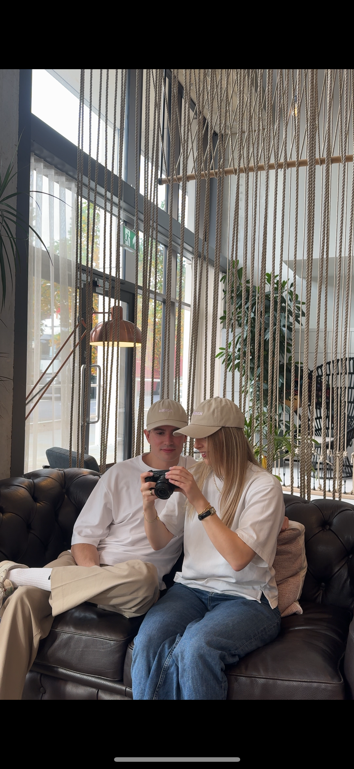 Two young people, one male and one female, sitting on a black leather couch in a modern cafe or lounge, looking at a camera held by the female. Both are wearing beige caps and casual clothing, with the female showing a friendly expression as she shows the camera to the male.