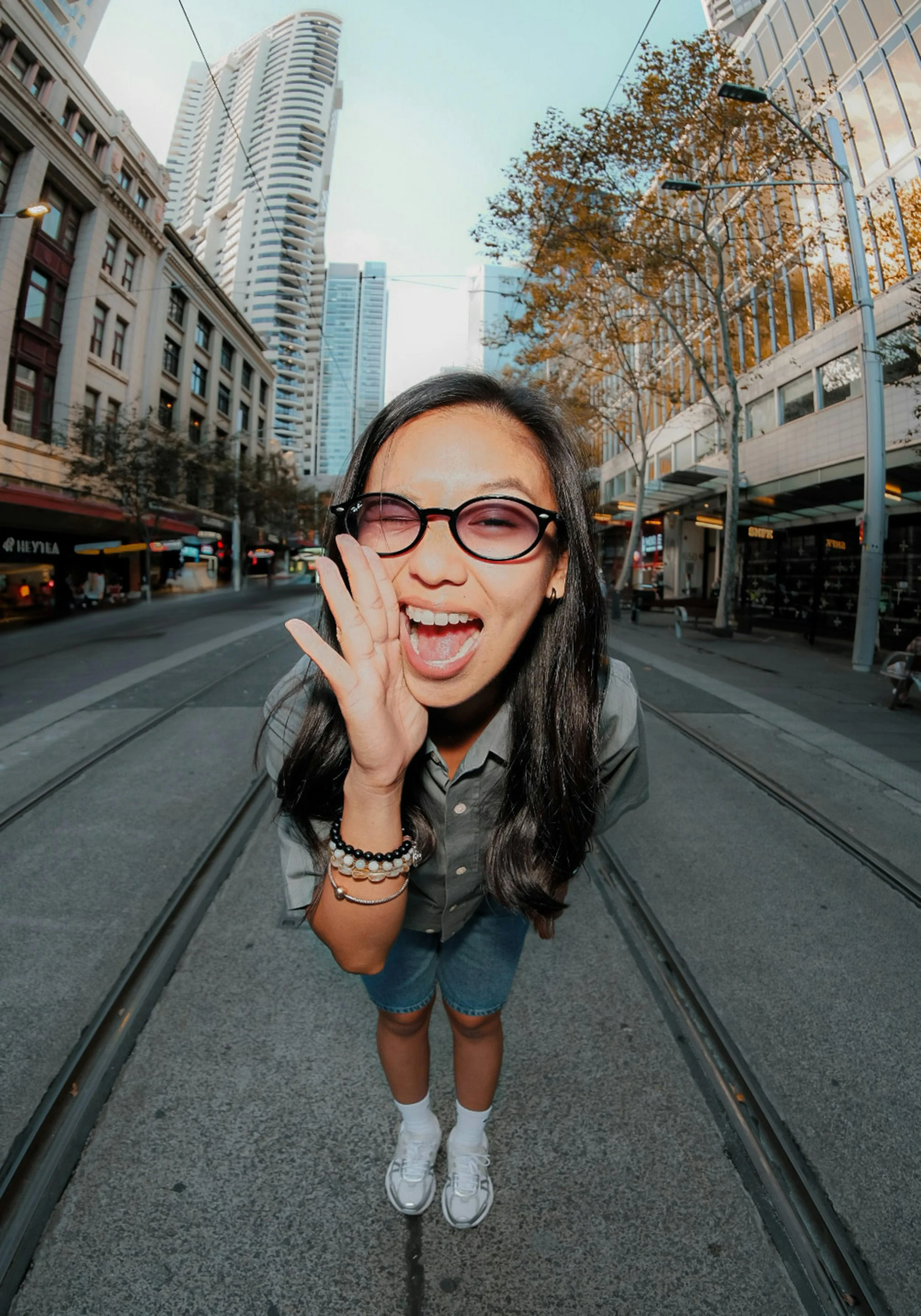 Young woman with glasses, smiling and cupping her ear, standing on tram tracks in a city street with tall buildings and trees.