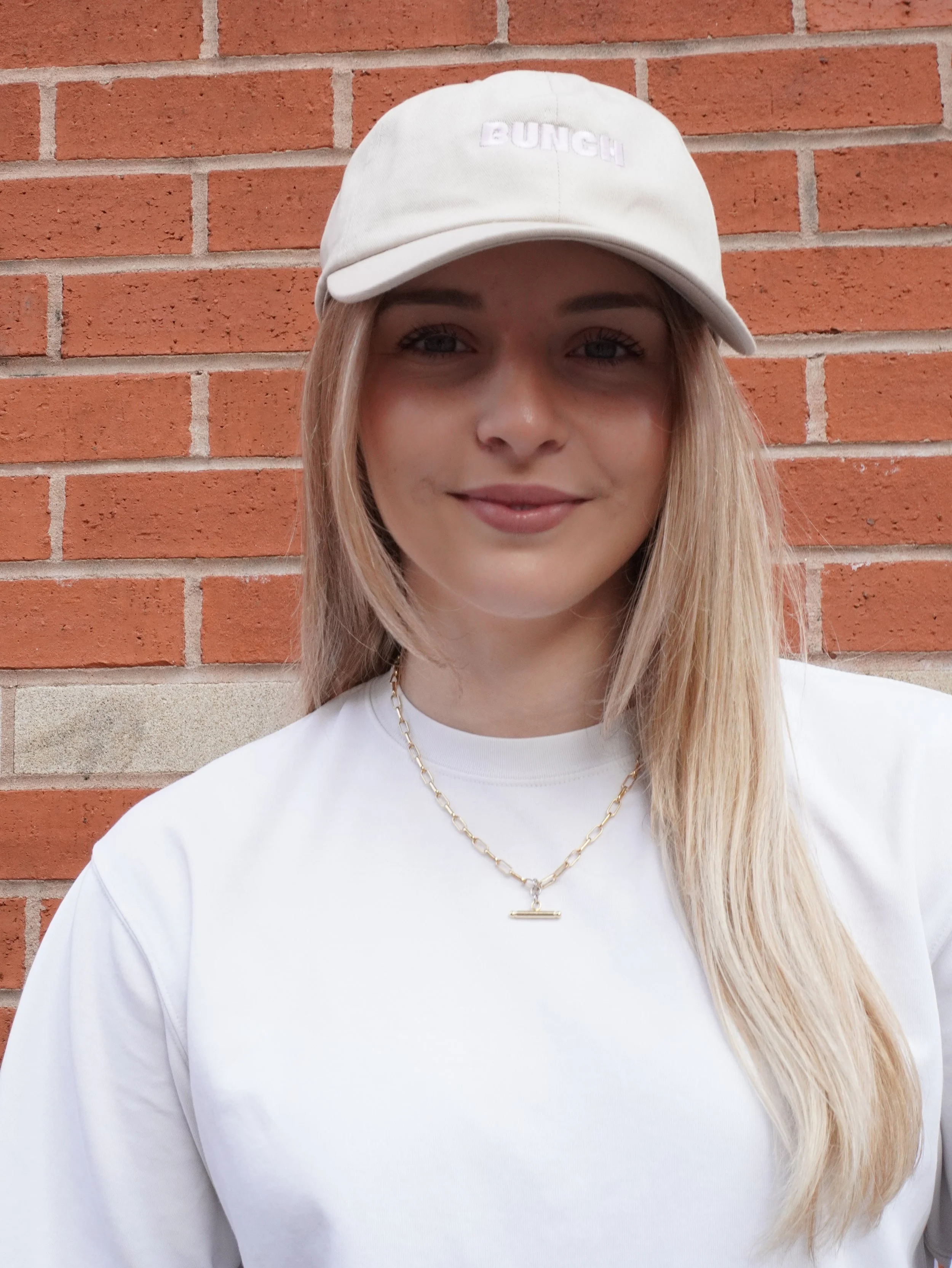 A young woman with long blonde hair, wearing a cream-colored baseball cap with 'BUNCH' embroidered on it, a white t-shirt, and a gold chain necklace, standing in front of a brick wall.