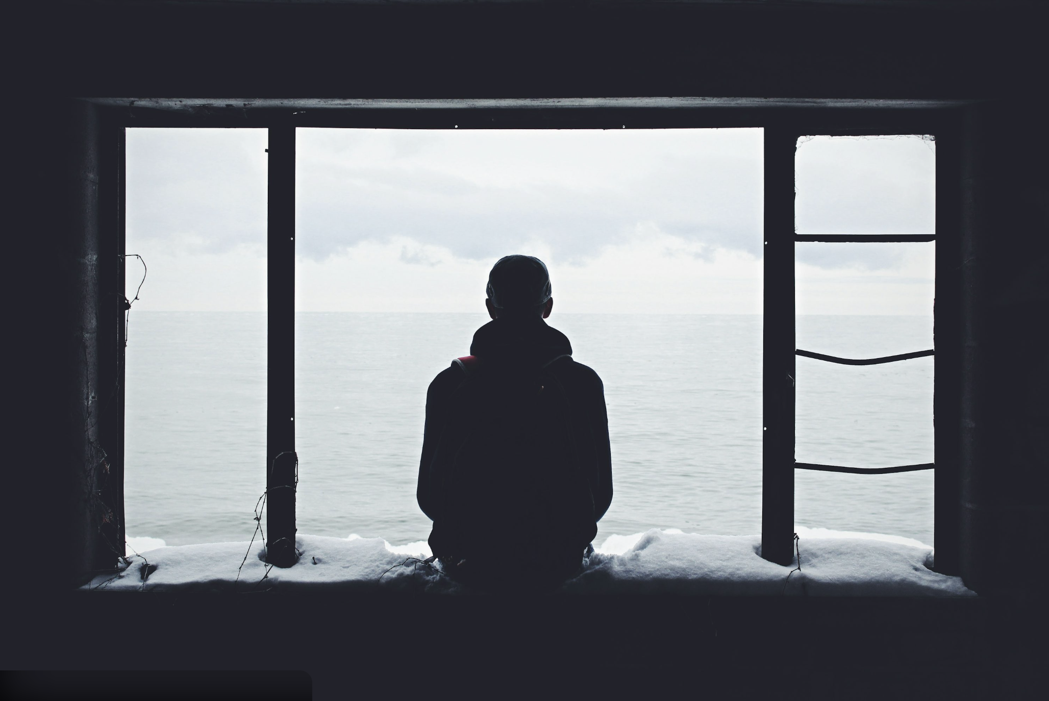 Man sitting quietly indoors, looking out at the ocean, reflecting emotional distance and inner struggle.