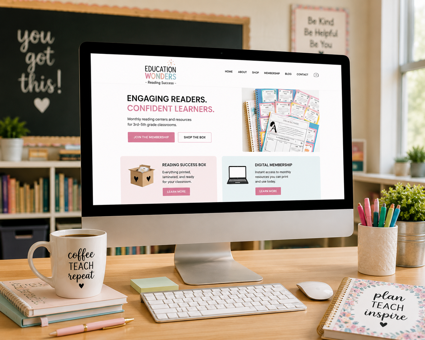 Computer monitor displaying an educational website about reading resources, surrounded by a coffee mug with motivational words, notebooks, sticky notes, pens, and potted plants on a wooden desk in a bright classroom setting.