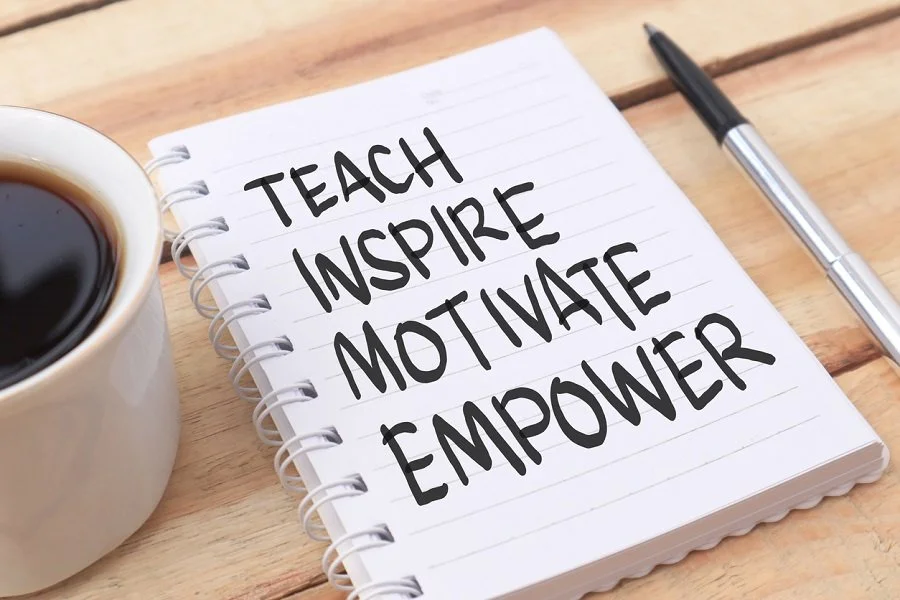 Notepad with the words 'Teach Inspire Motivate Empower' written on it, with a pen and a cup of coffee on a wooden table.