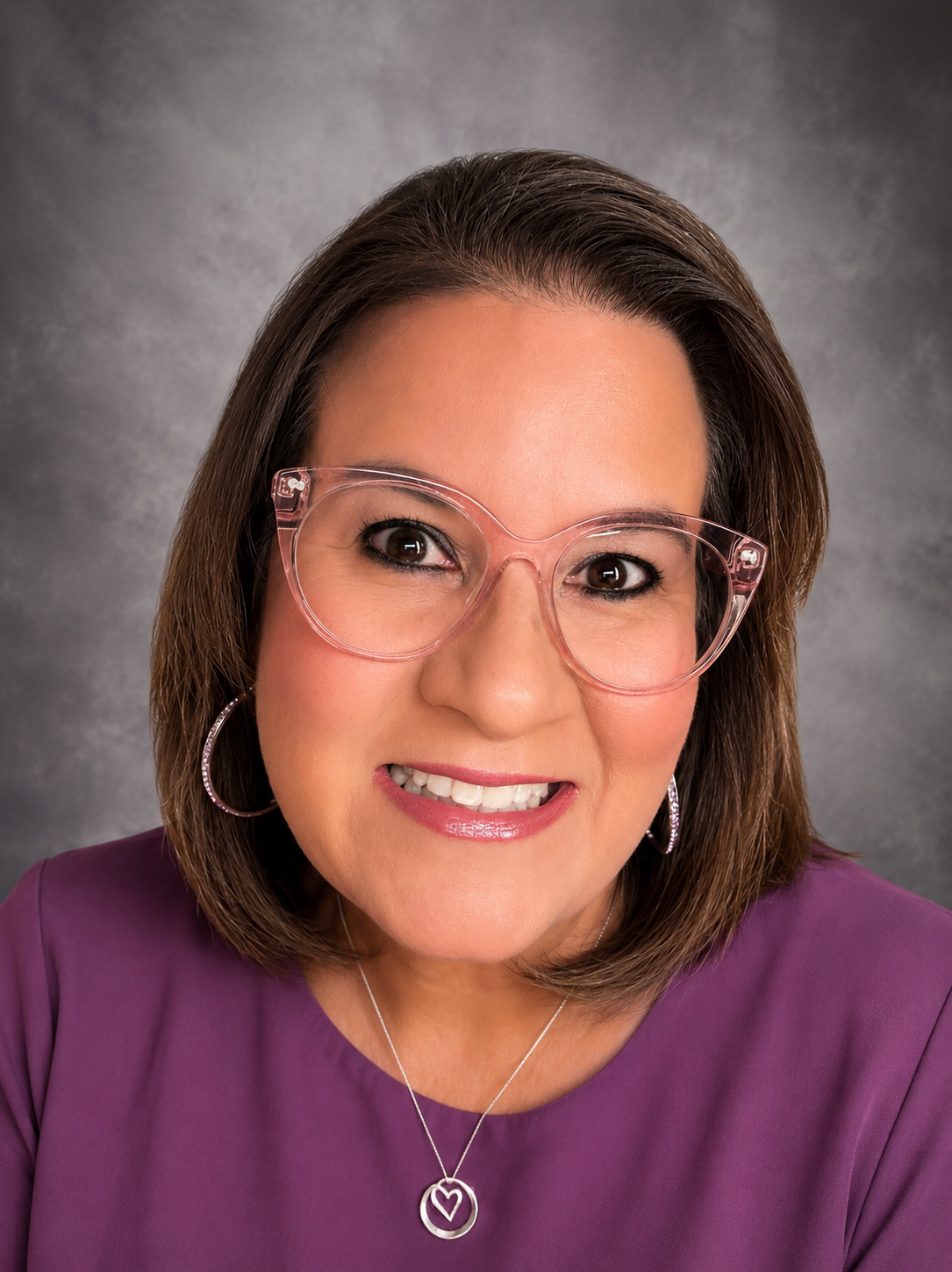 A woman with shoulder-length brown hair wearing large pink glasses, hoop earrings, a purple top, and a silver necklace with a heart pendant, smiling at the camera against a gray textured background.