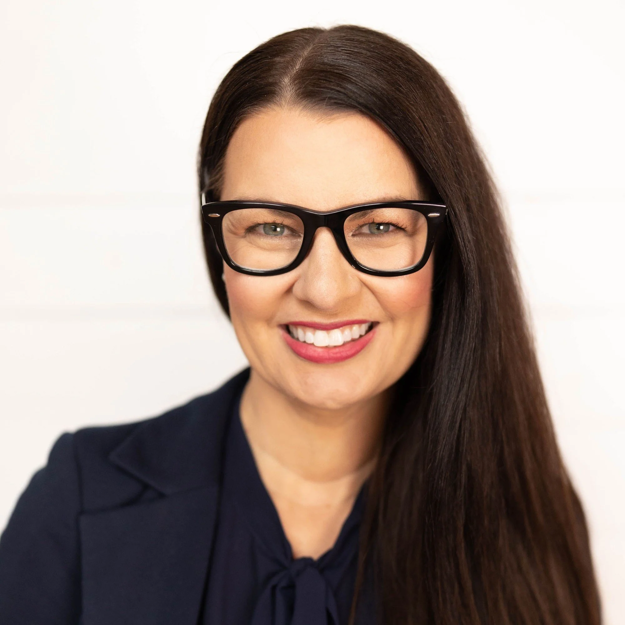 Close-up of a smiling woman with long dark hair, wearing black glasses and a navy blazer, posed against a white background.