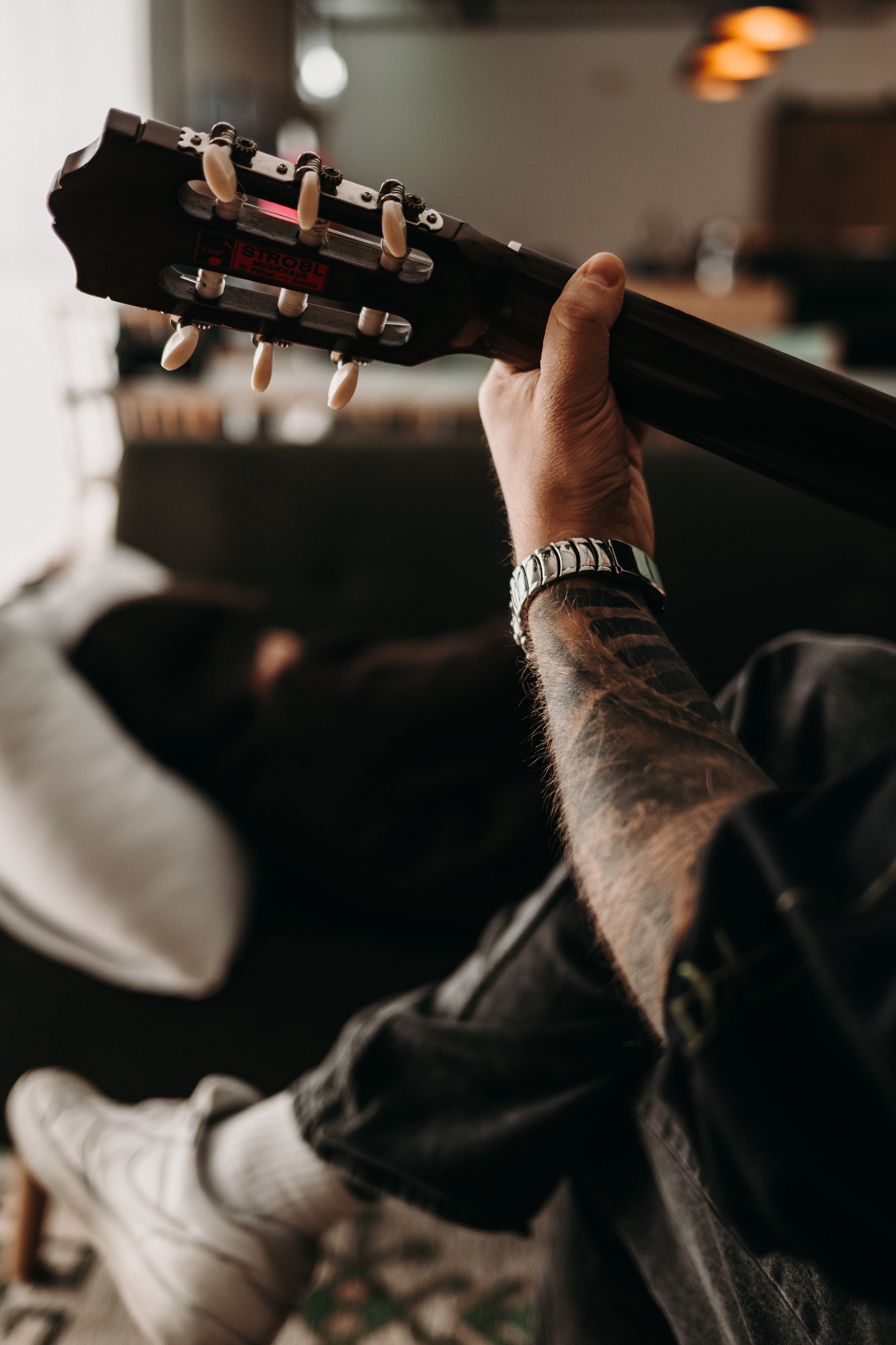A person with a tattooed arm and a silver watch playing an acoustic guitar indoors, sitting on a couch.