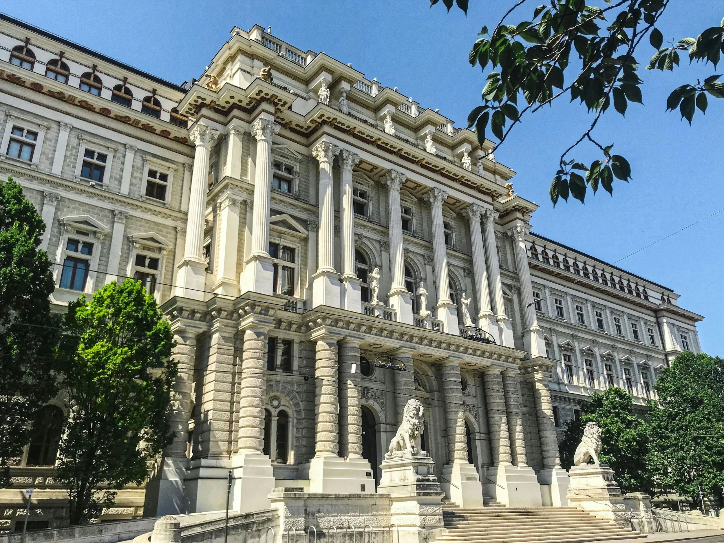 Una grandiosa edificación histórica de estilo neoclásico, con columnas dóricas y estatuas en la entrada, rodeada de árboles y un cielo azul claro.
