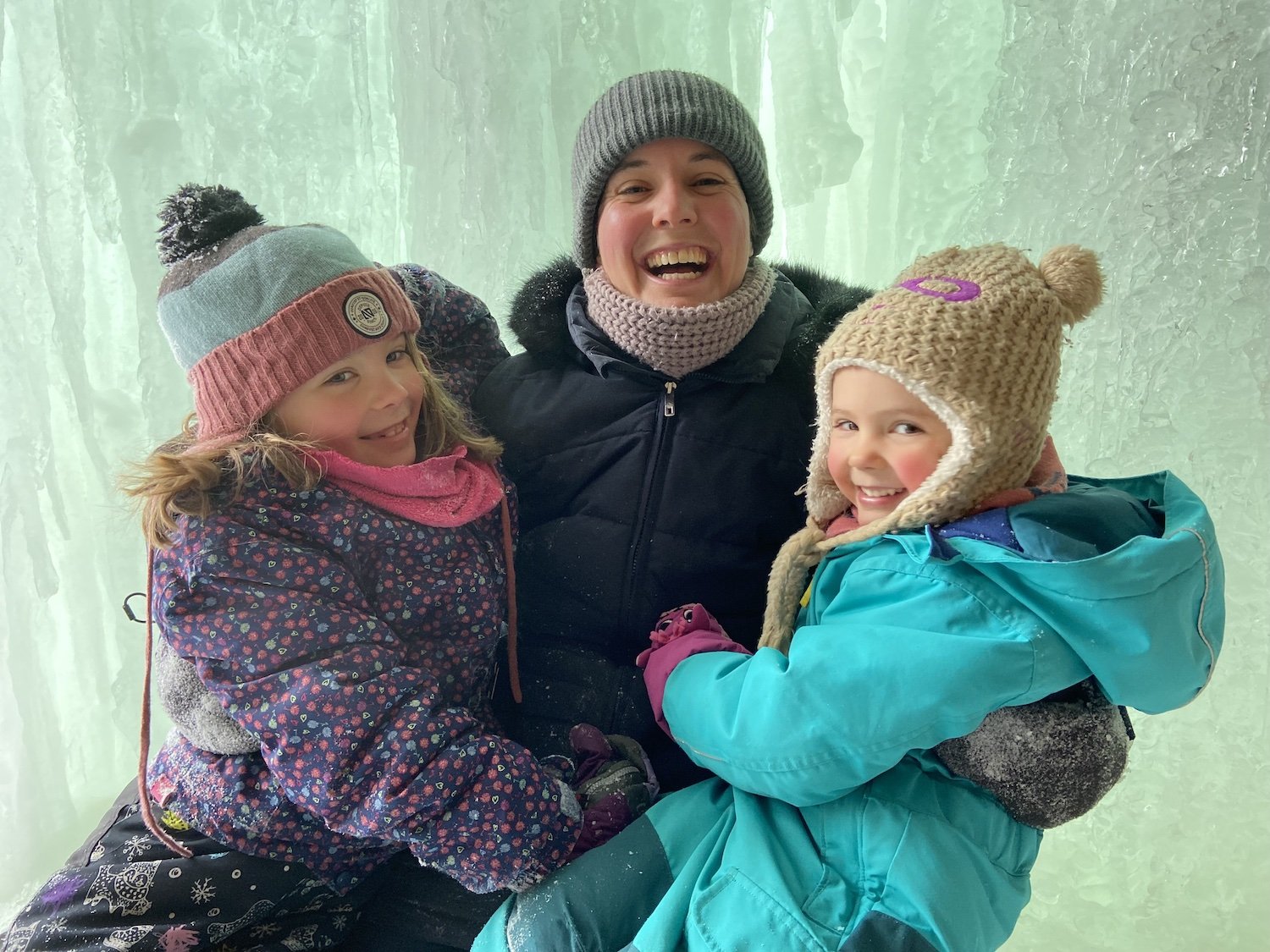 A smiling woman in winter clothing, including a gray beanie and scarf, poses with two young girls in colorful winter jackets and hats inside an ice cave with translucent ice walls.