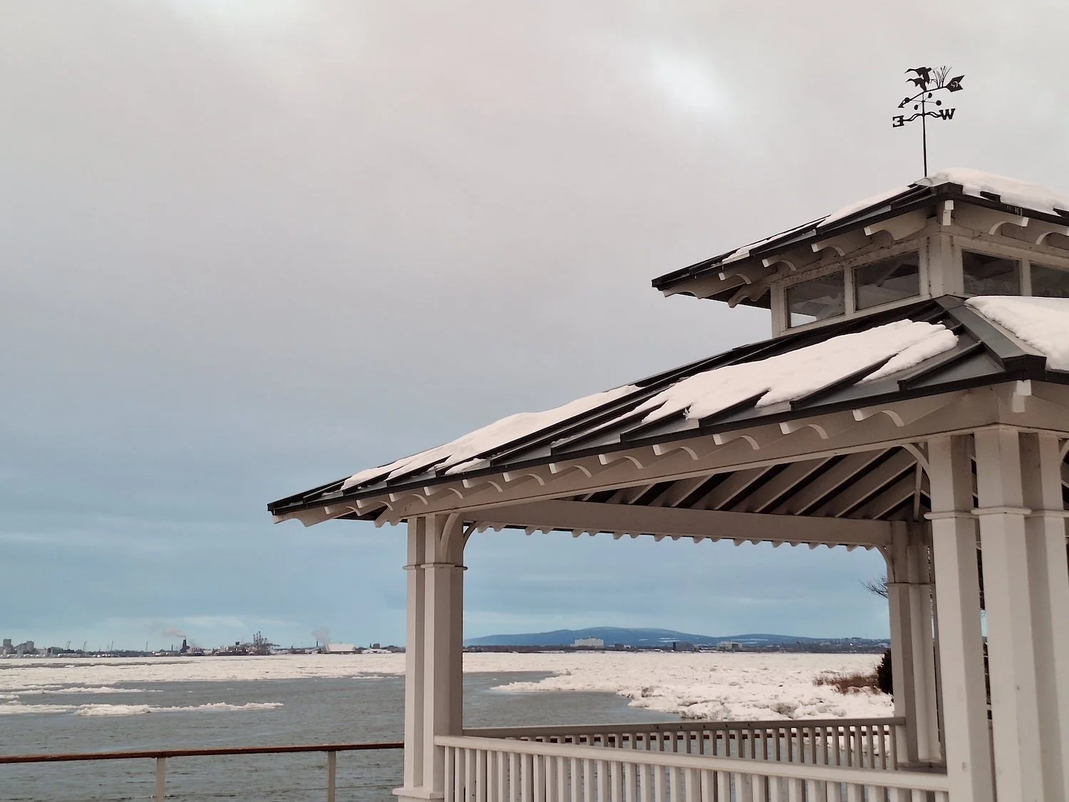 A white beach hut with snow on its roof, overlooking a frozen body of water with distant industrial buildings and mountains in the background.
