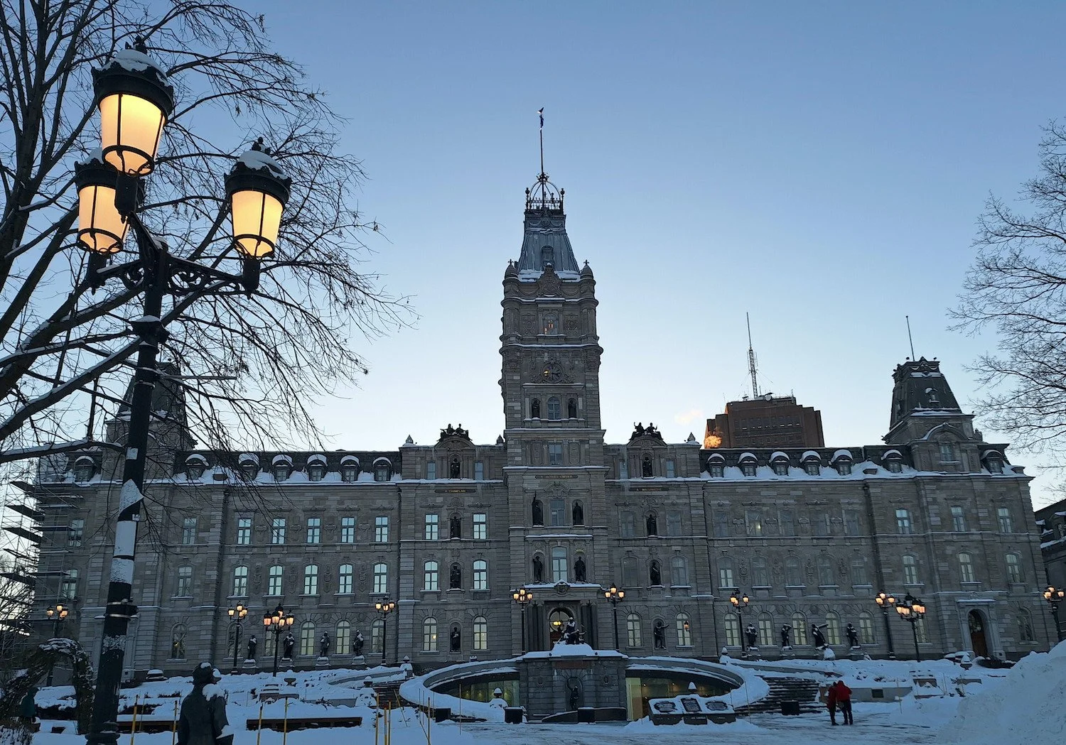 Historical government building with a central clock tower, snowy surroundings, leafless trees, and street lamps, under a clear sky.