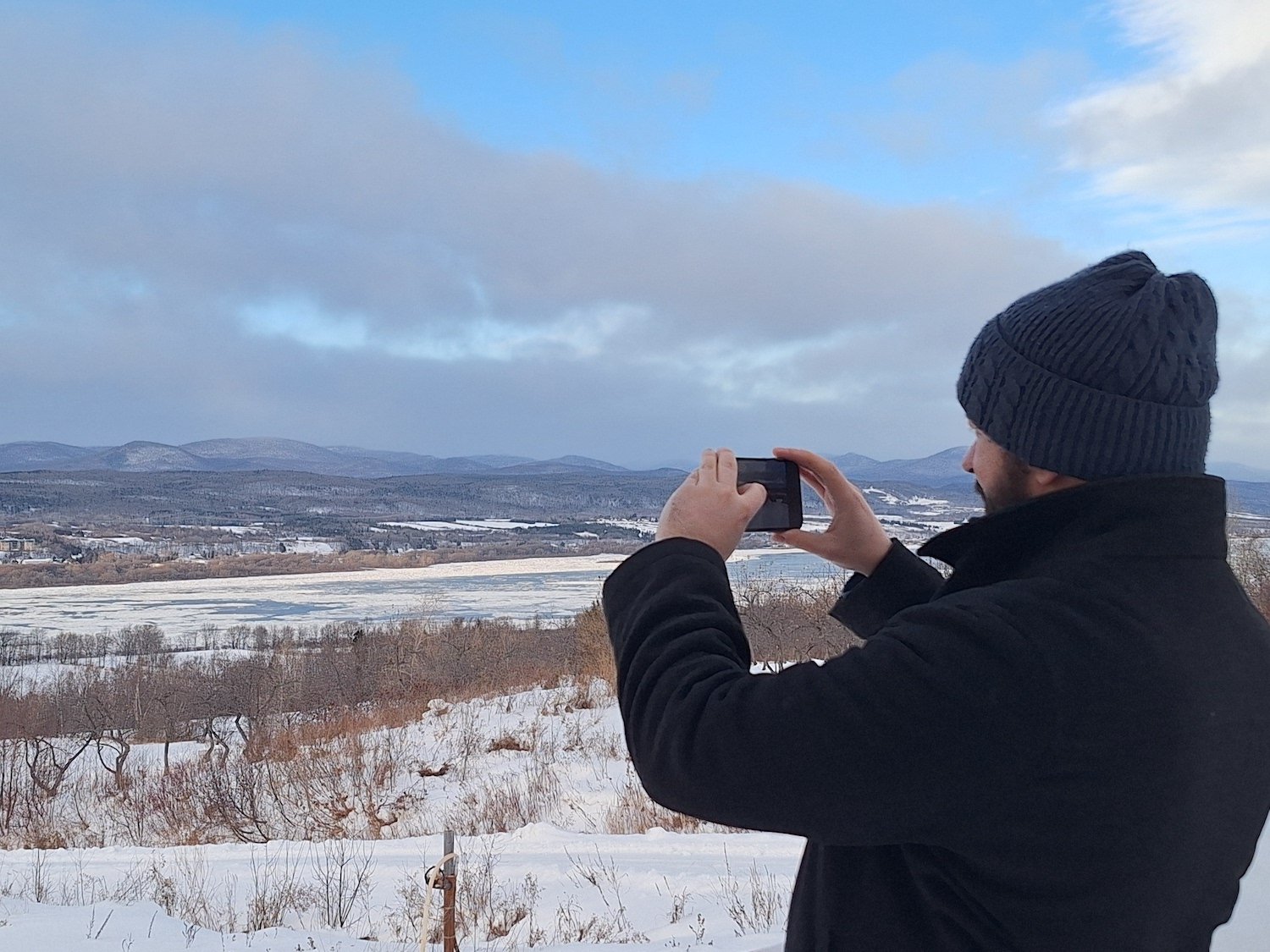 A man wearing a black jacket and a gray knit beanie taking a photo with his smartphone of a snowy winter landscape with hills and a partly cloudy sky.