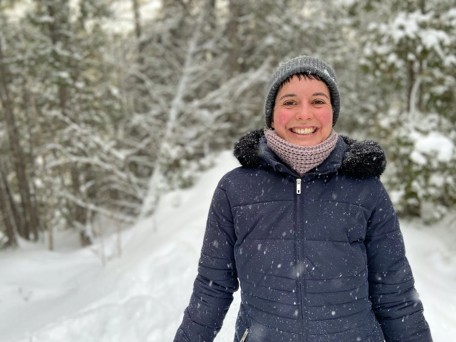 A woman smiling outdoors in a snowy landscape, wearing a dark blue winter coat, a gray knit hat, and a purple knit scarf.