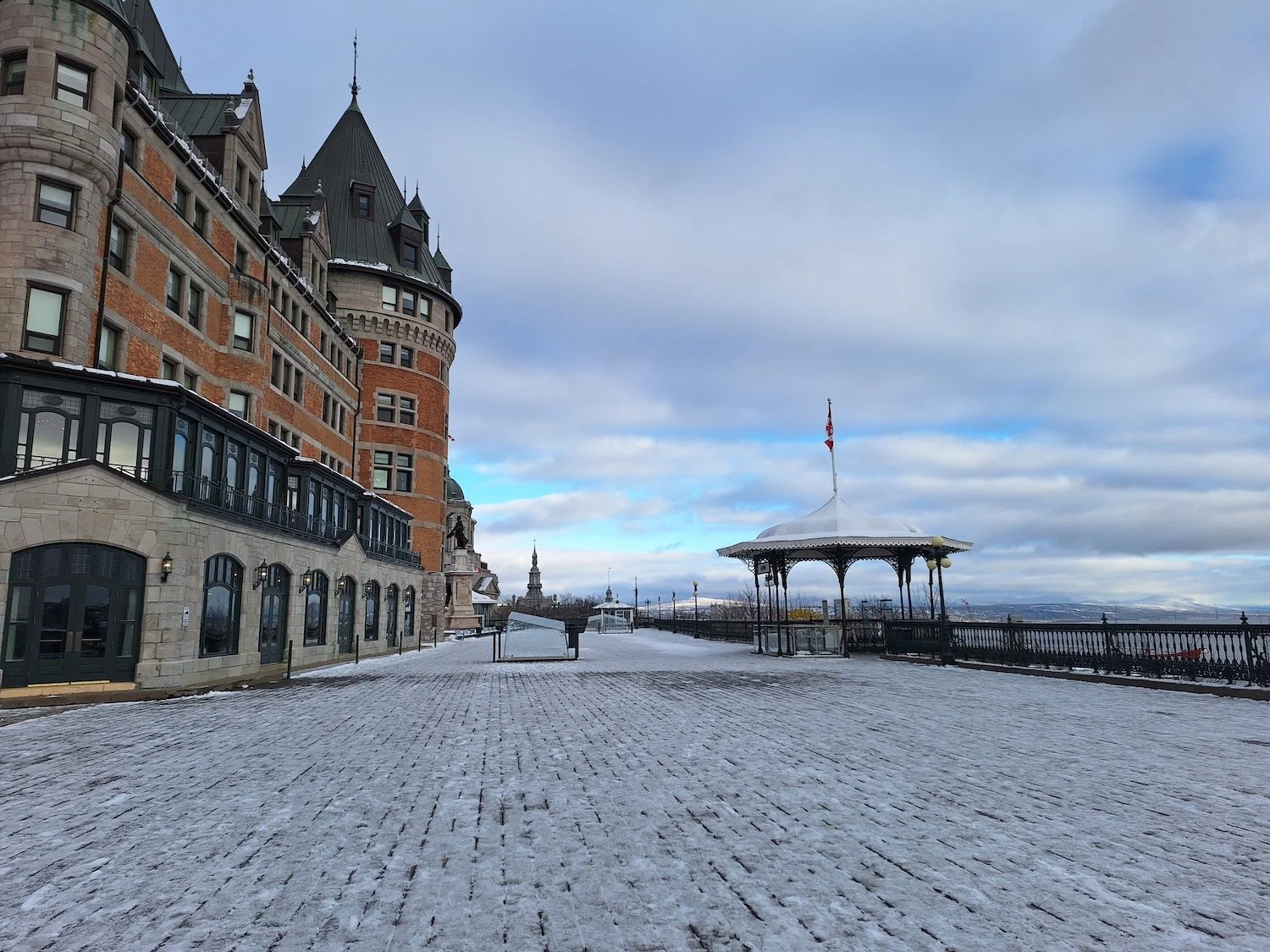 View of a historic building with turrets and a wraparound porch, a snow-covered promenade, and a gazebo with a Canadian flag on top, overlooking a cloudy sky.