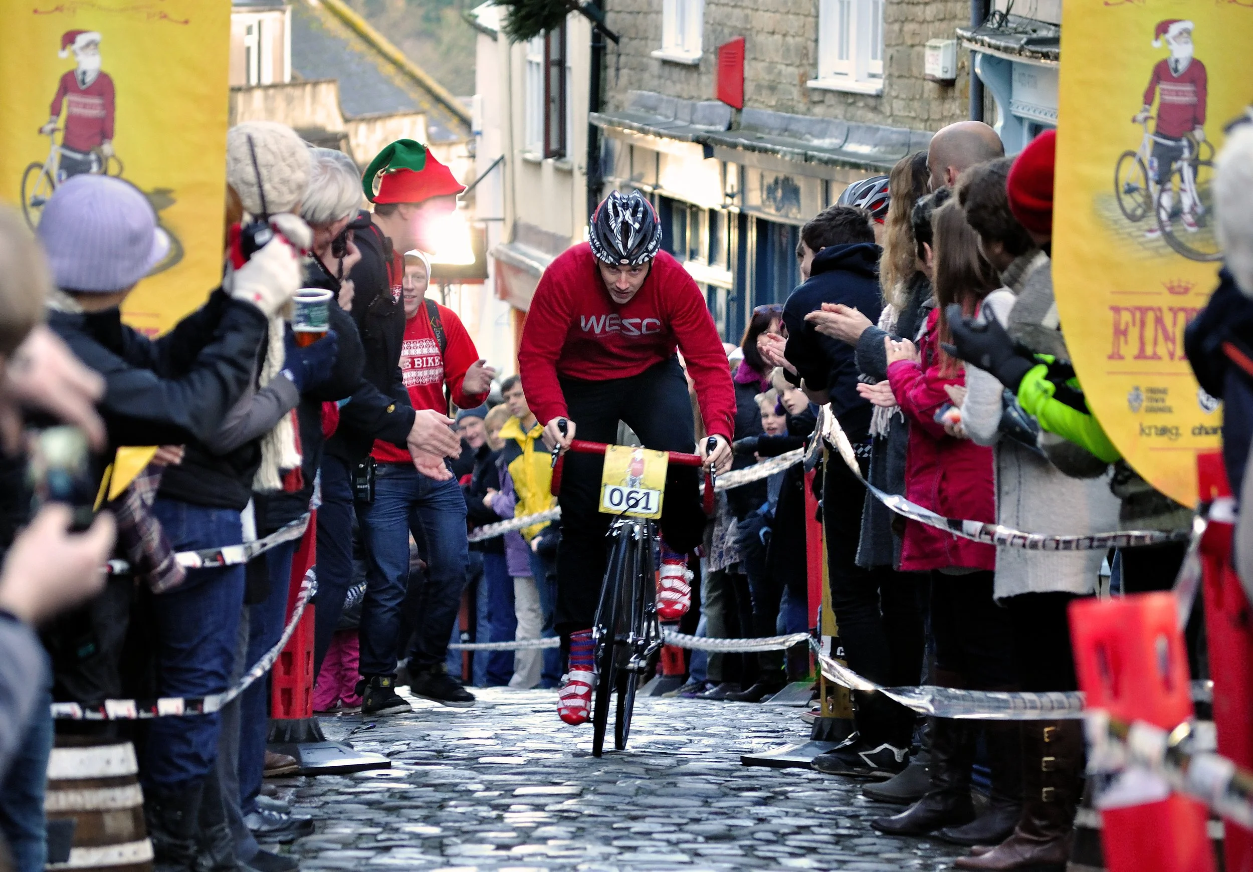 Cyclist riding a bike over a cobblestone street amidst a crowd of spectators on both sides, during a race event with banners.
