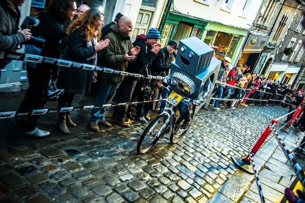 A person wearing a robot costume is riding a bicycle during a street event, with a group of spectators watching behind a barricade, on a wet cobblestone street.