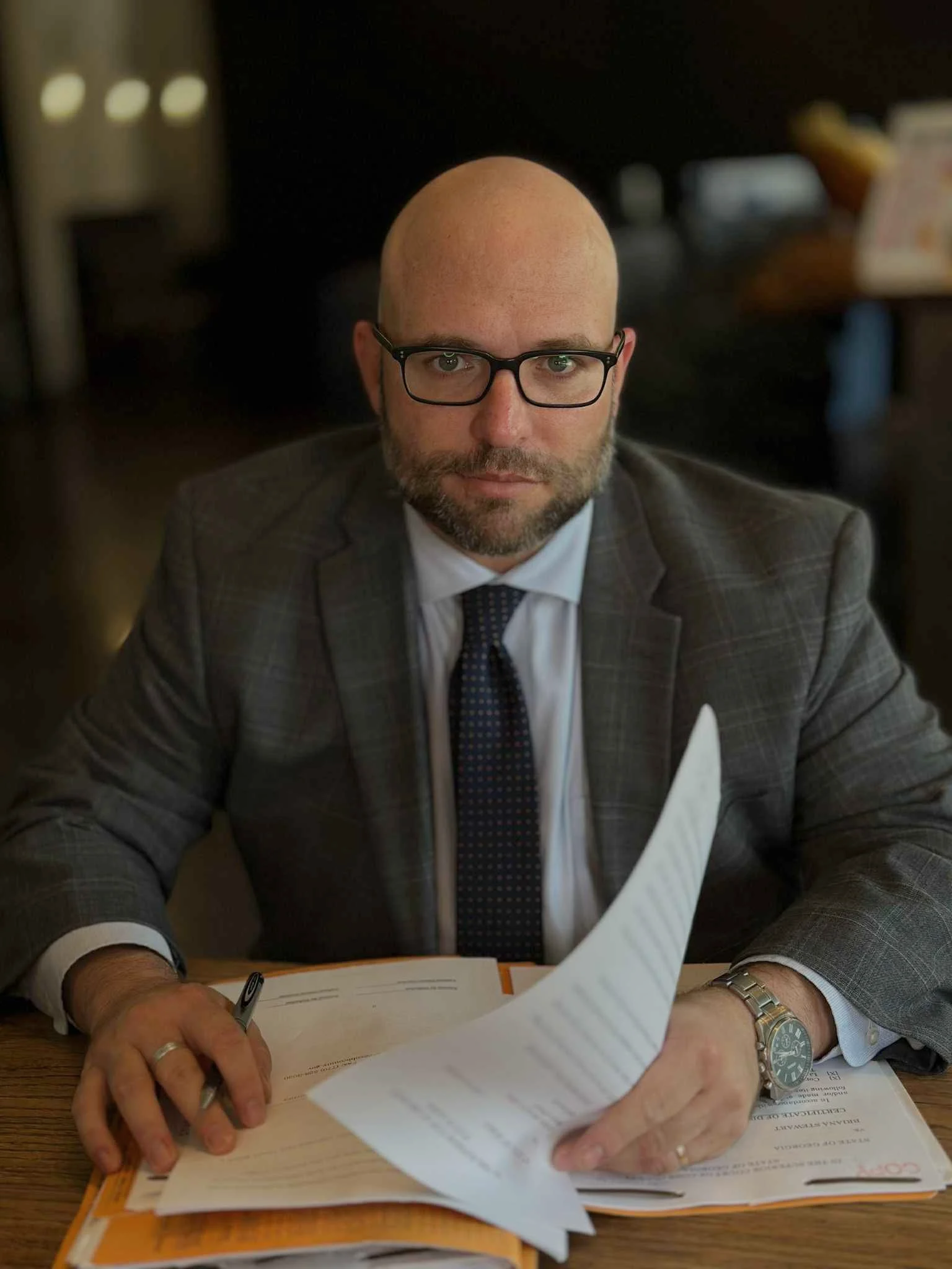 Jeremy Hager, the criminal defense attorney. He is dressed in a plaid suit, white shirt, black tie, and glasses. He sits at his office desk with documents, holding a pen in one hand and turning a page with the other.