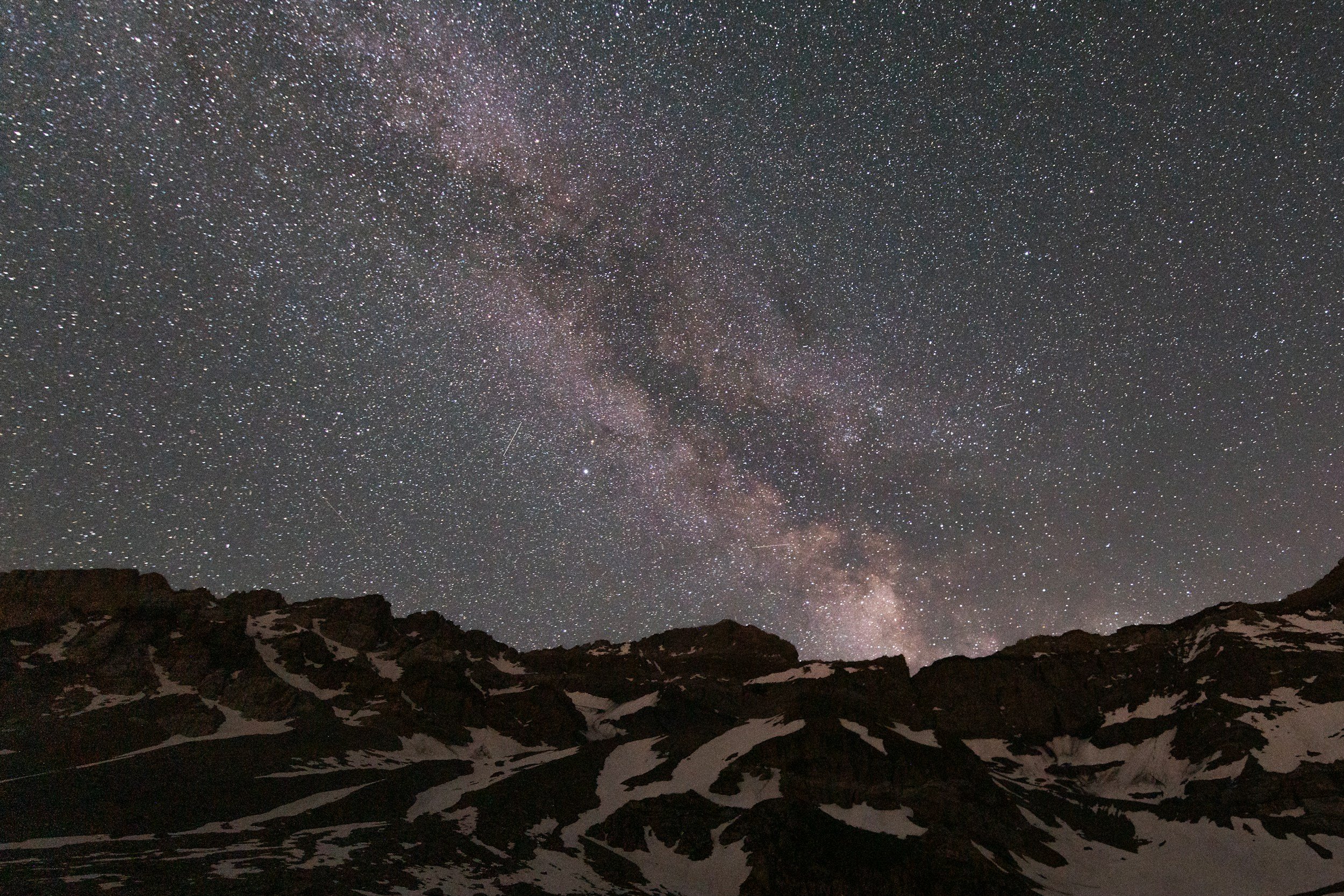 Night sky filled with stars and the Milky Way galaxy above snow-capped mountain peaks.