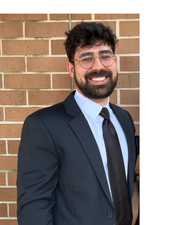 A young man with dark curly hair, glasses, and a beard, wearing a black suit, white dress shirt, and black tie, smiling and standing against a brick wall.