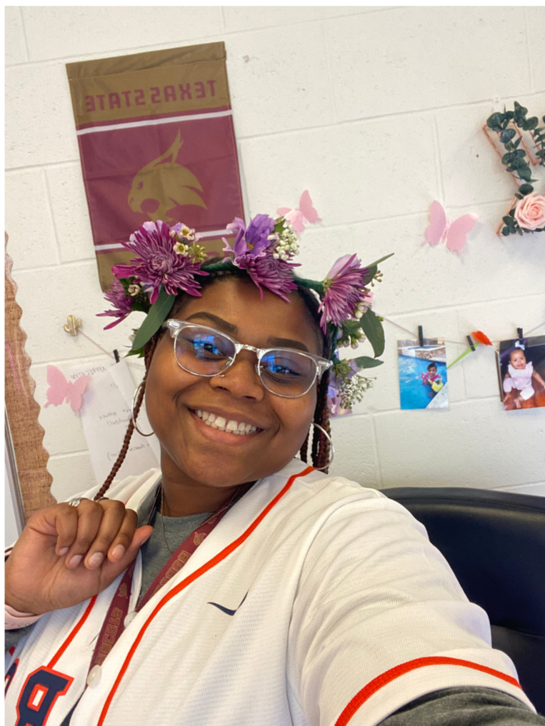 A smiling woman wearing glasses and a white sports jersey with orange and navy accents, accessorized with a flower crown made of purple flowers and greenery. The background shows a wall decorated with pink butterfly cutouts, small photographs, and a Texas State flag.