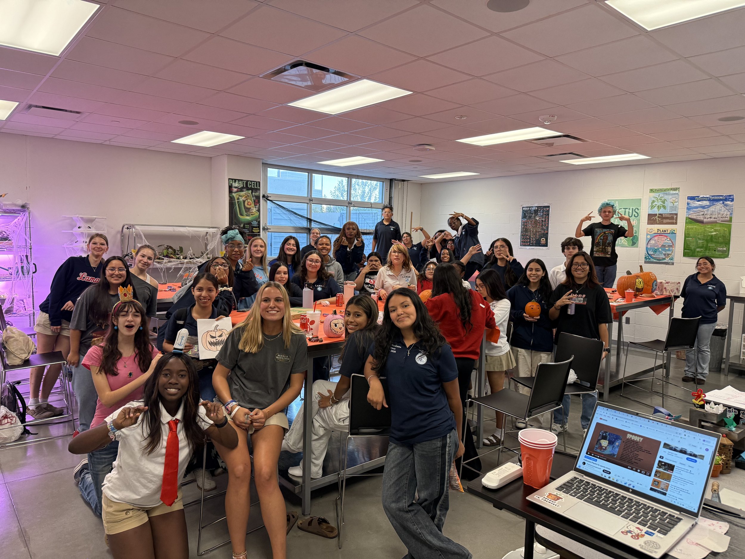 Group of students in a classroom decorated with Halloween and plant-themed decorations, some dressed in costumes, engaged in a Halloween activity.
