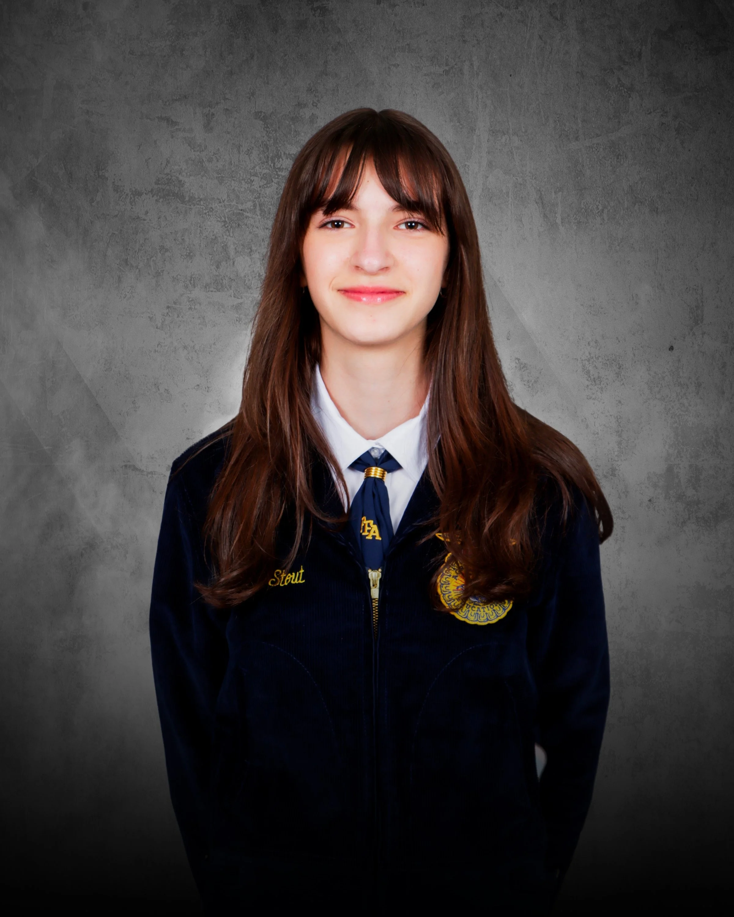 A young woman with long brown hair, wearing a navy blue jacket, white shirt, and a blue tie, standing against a beige stone wall, smiling at the camera.