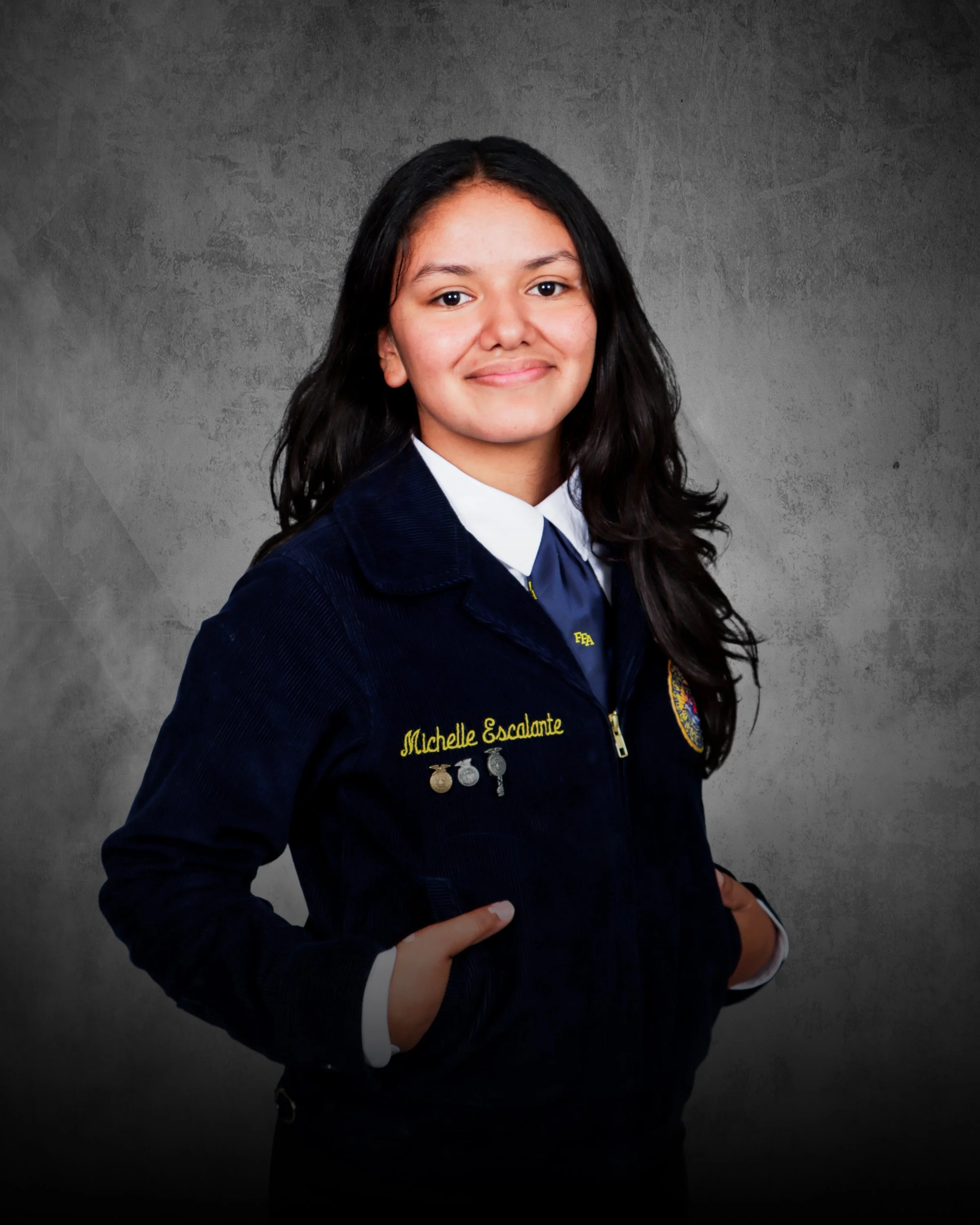 Young girl with long dark wavy hair smiling, wearing a navy zipper jacket with a white shirt and navy tie, standing against a beige brick wall.