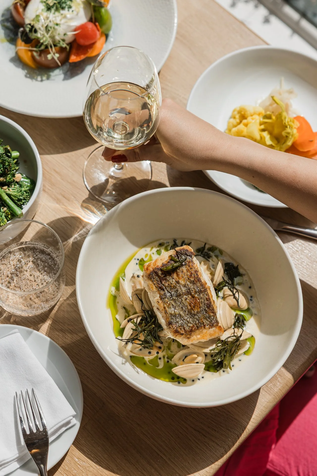 A meal with a piece of grilled fish on top of a bed of pasta and garnished with herbs and black sesame seeds, served with green oil or sauce, on a white bowl. Surrounding it are other plates with salads and wine glasses with white wine.