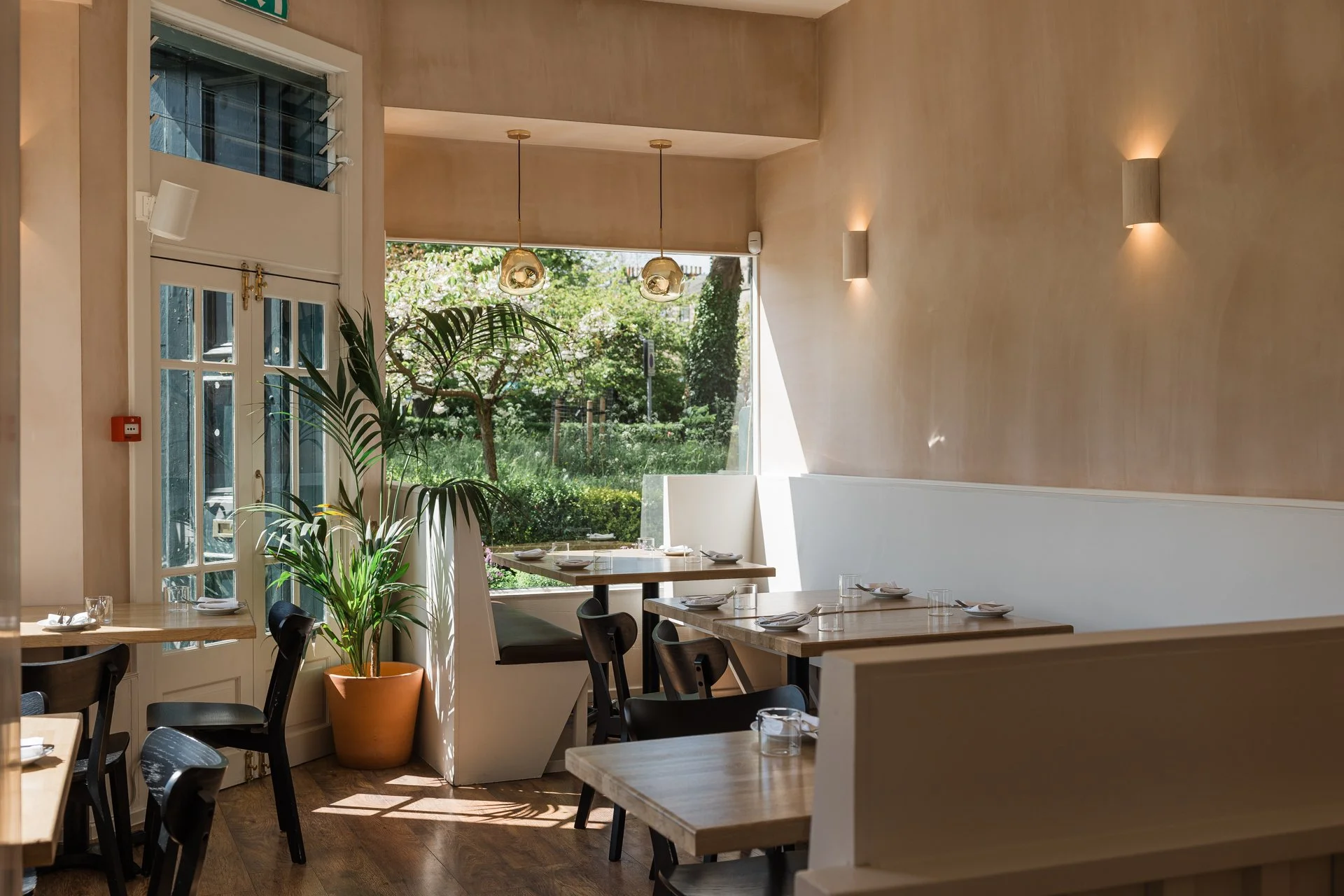 Interior of a bright, modern restaurant with wooden tables, black chairs, wall-mounted lights, and large windows showing greenery outside.
