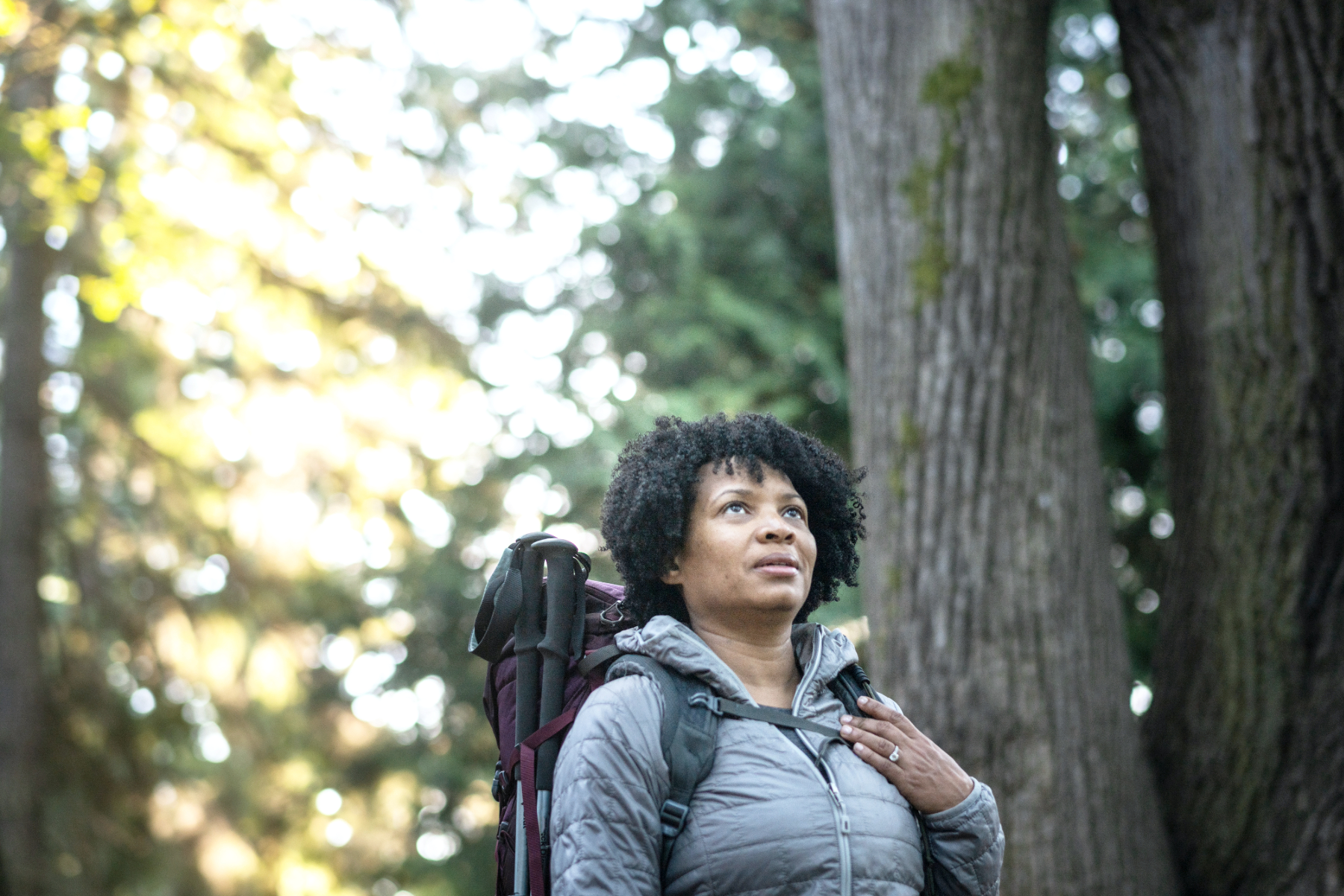 Woman with a backpack in a forest, looking upwards with a thoughtful expression.